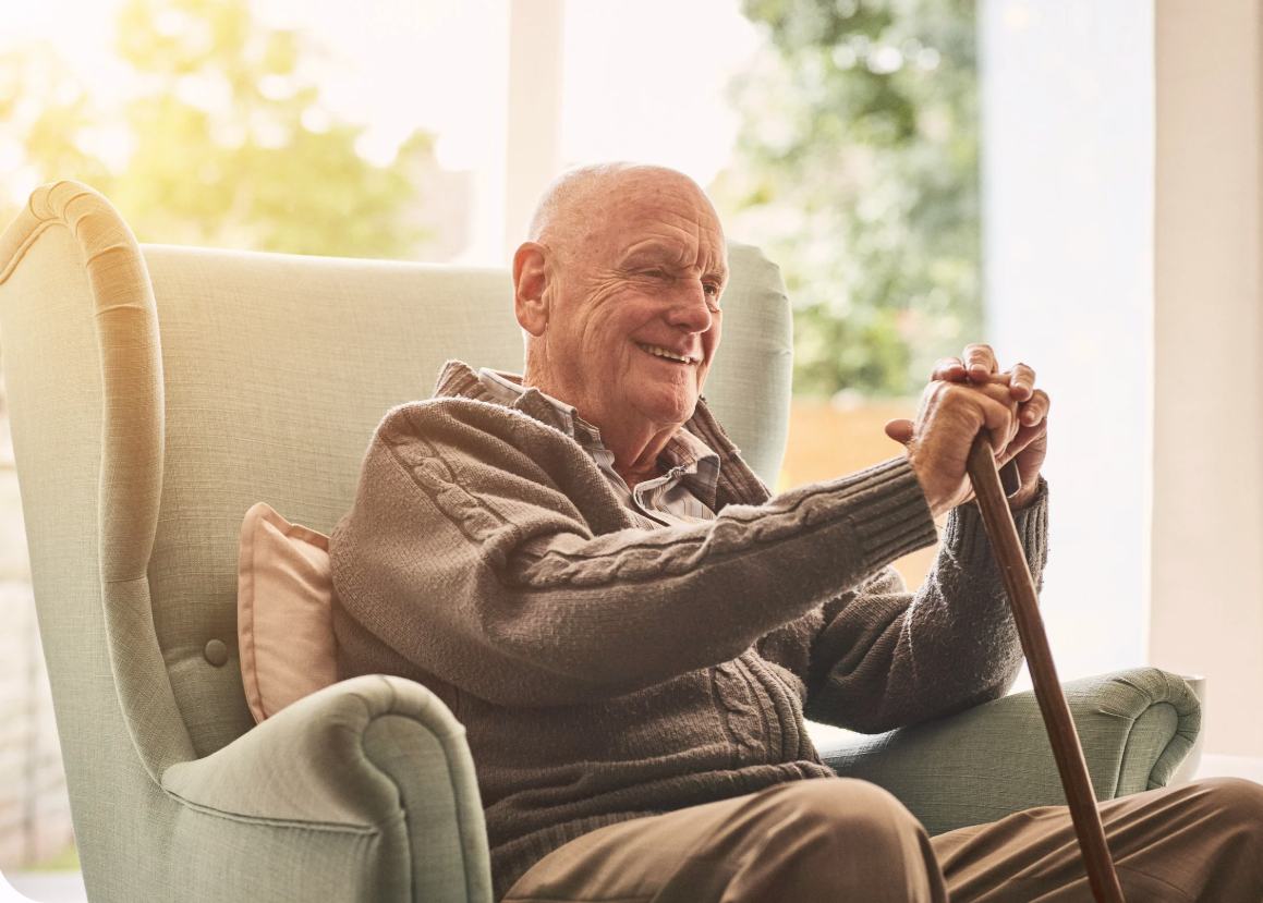 An elderly man sitting in a comfortable armchair, smiling, with a walking cane in his hands. The room is bright with sunlight and a view of trees outside.