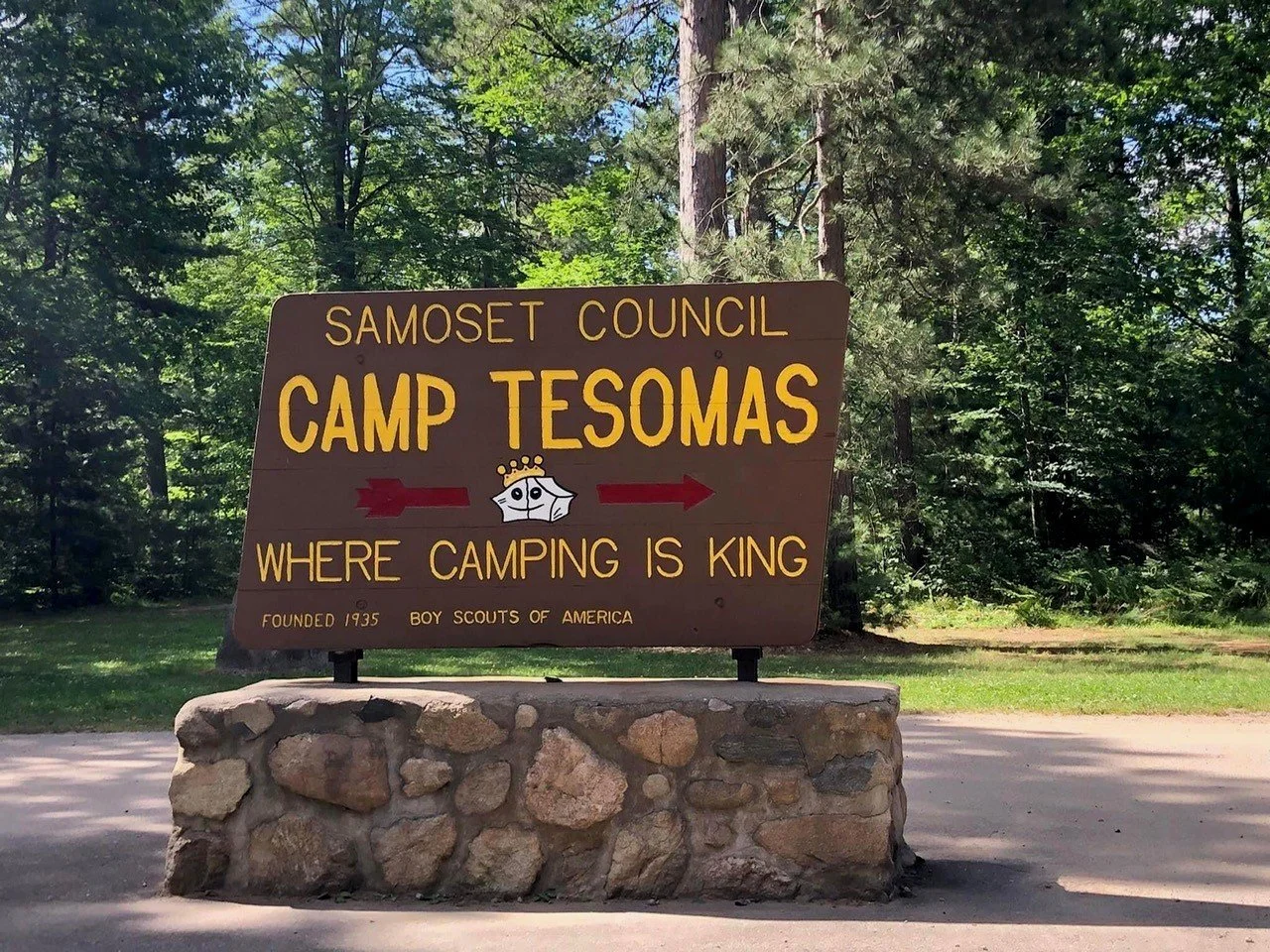 Sign for Camp Tesomas, by Samoset Council, with a small cartoon crown and arrow pointing right, surrounded by trees in a wooded area.