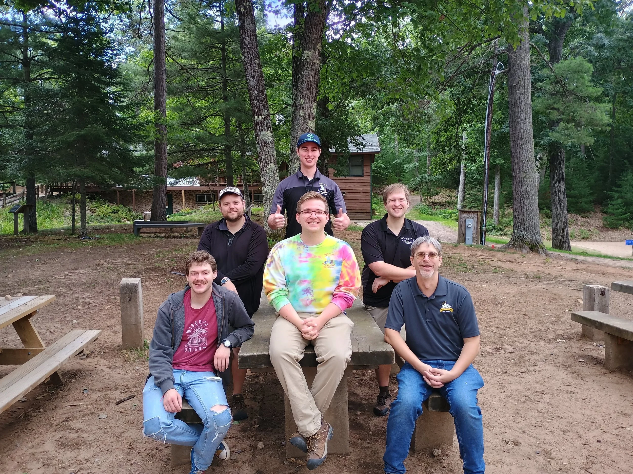A group of nine young adults outdoors in a wooded area, posing for a photo with some sitting on benches and others standing behind them, smiling and appearing happy.