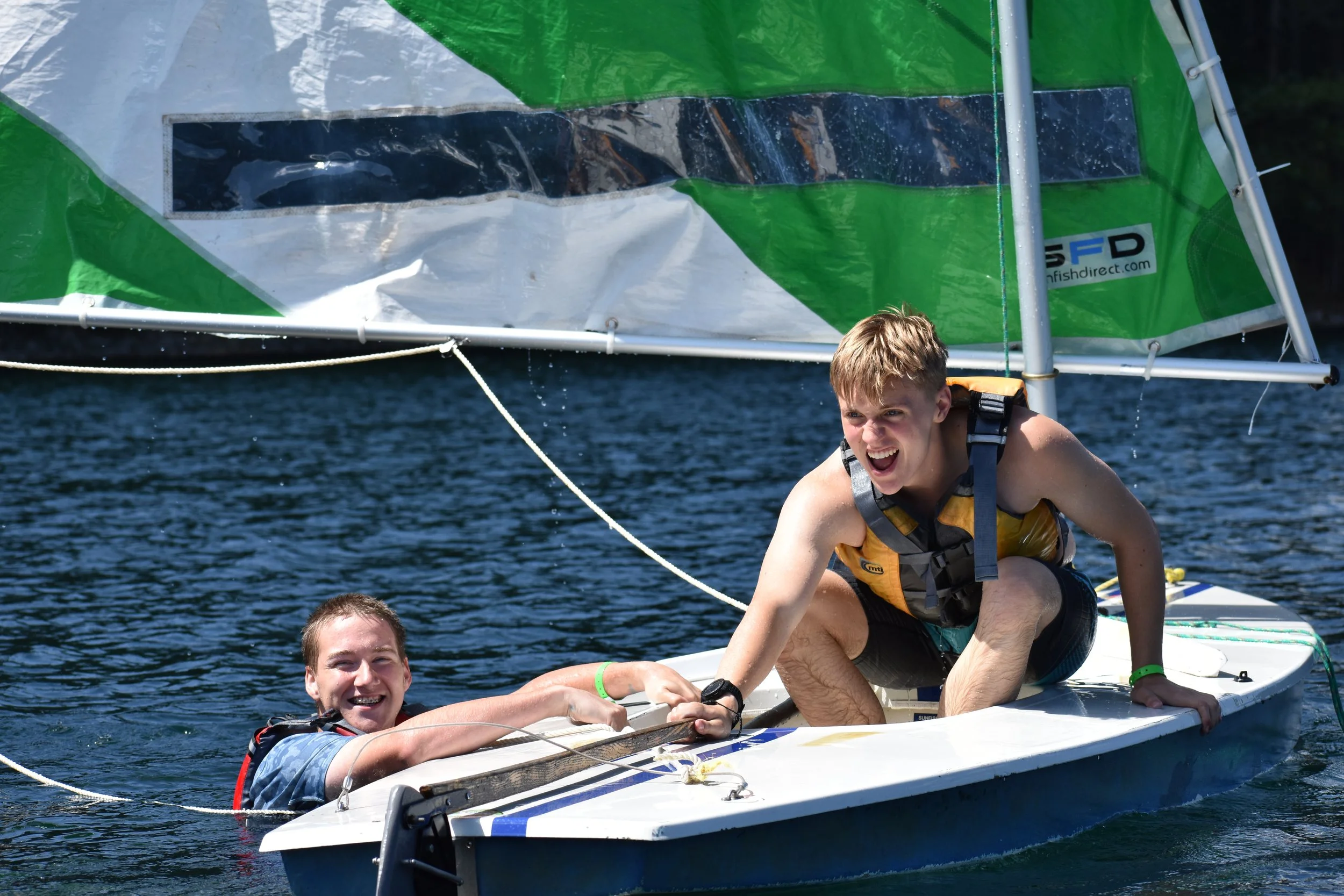 Two young men smiling on a small sailboat on the water during daytime, with a sail and water visible in the background.