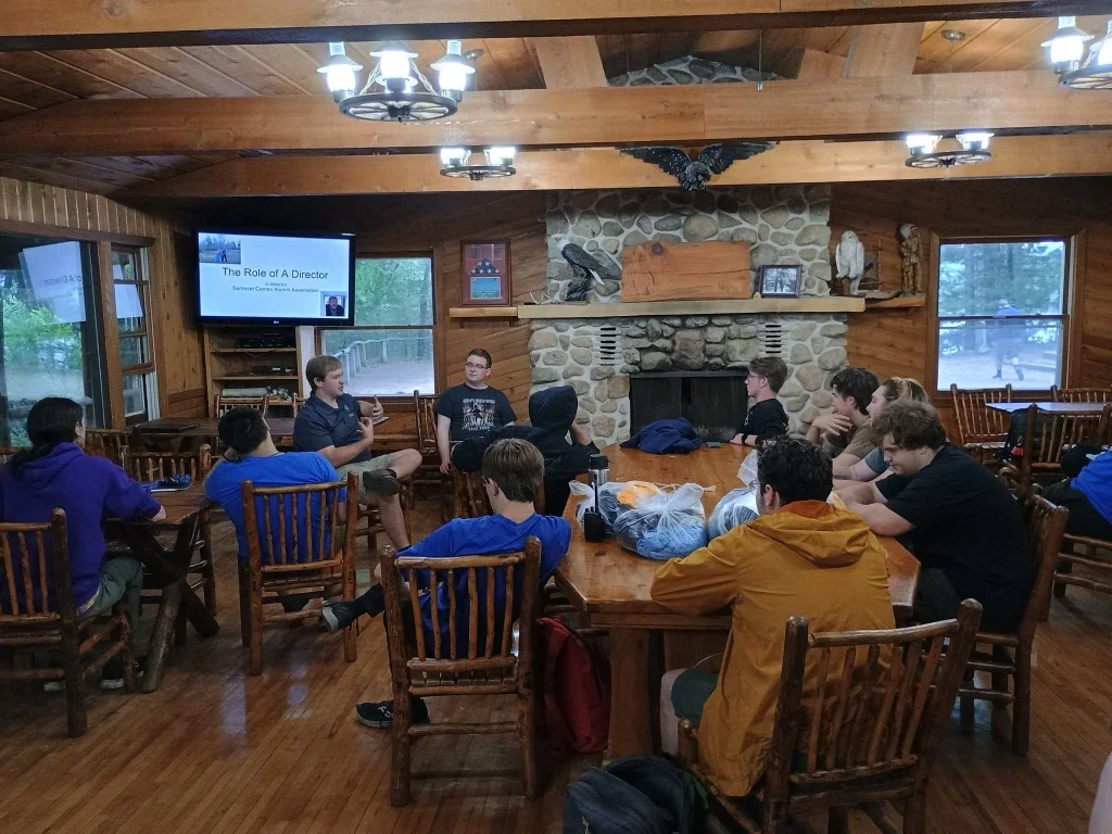Young people attending a presentation in a wooden cabin, seated around a large round table, with a fireplace and windows in the background.