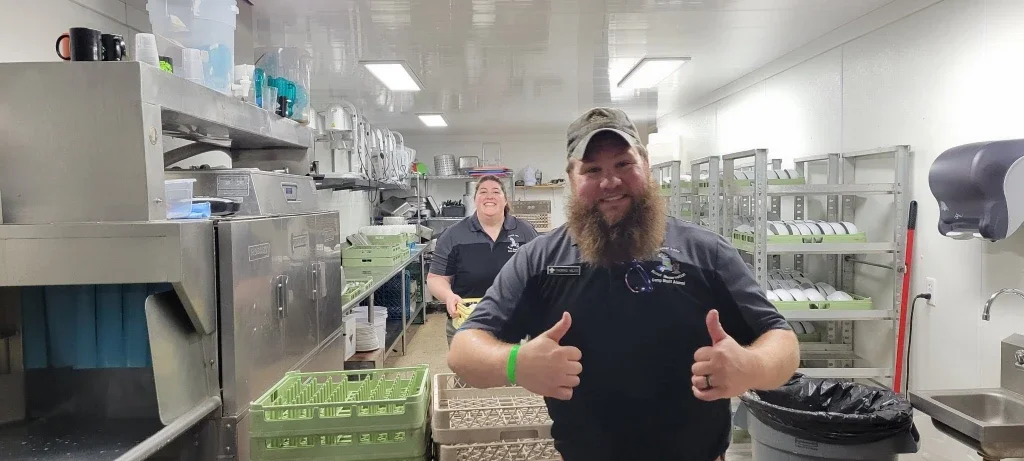 Two smiling kitchen staff in a commercial kitchen, giving thumbs up.