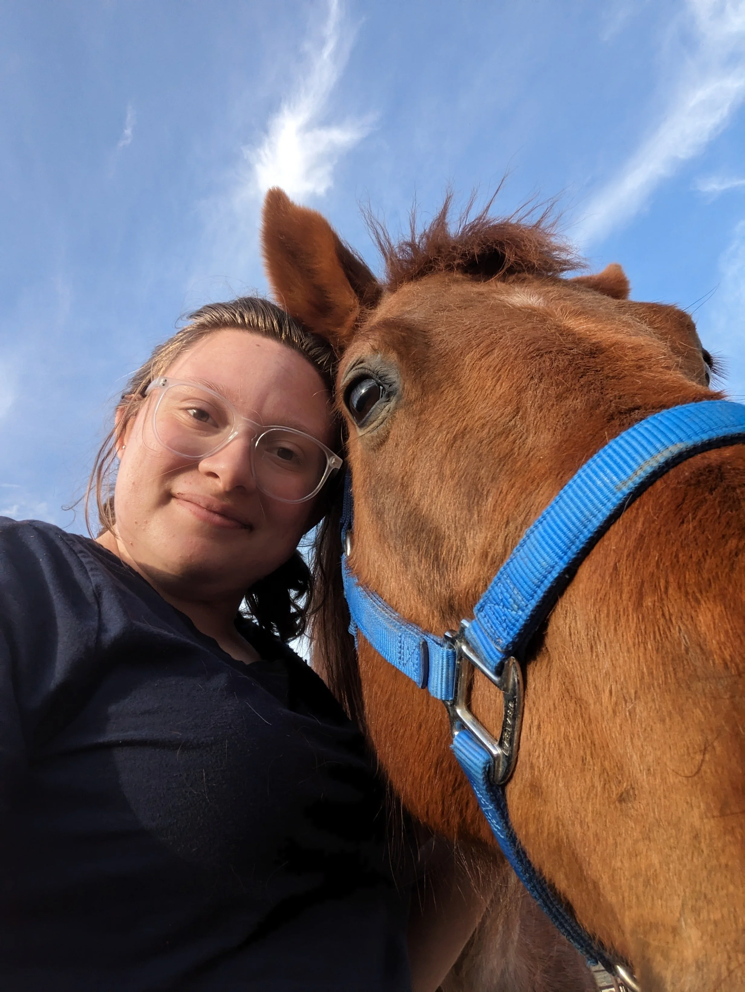 Close-up of a woman wearing glasses, smiling, with her face close to a horse's face against a blue sky with clouds.