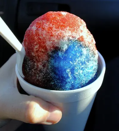 A hand holding a white bowl with a colorful snow cone in red, blue, and white ice shavings, with a white straw.