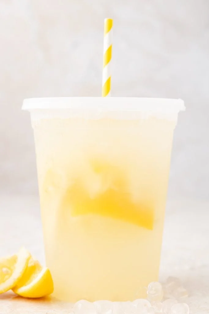 A clear plastic cup of lemonade with lemon slices and ice, with a yellow and white striped straw, placed on a light surface with lemon wedges and ice around it.