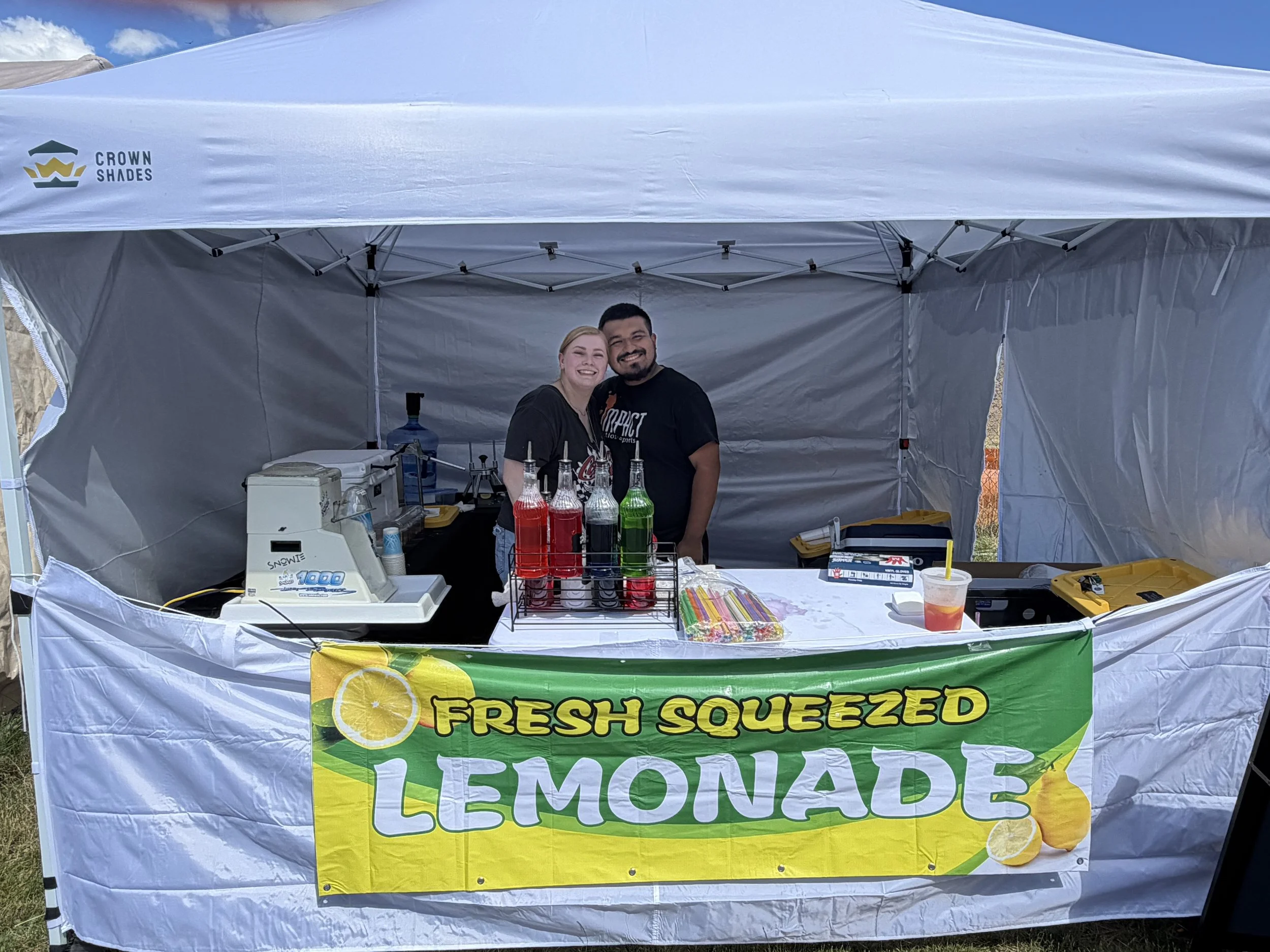 A lemonade stand under a white canopy tent with a yellow banner displaying the words 'Fresh Squeezed Lemonade' and images of lemons. Two smiling people, a woman and a man, stand behind the stand, which has bottles of colorful liquids, a cash register, and other supplies.