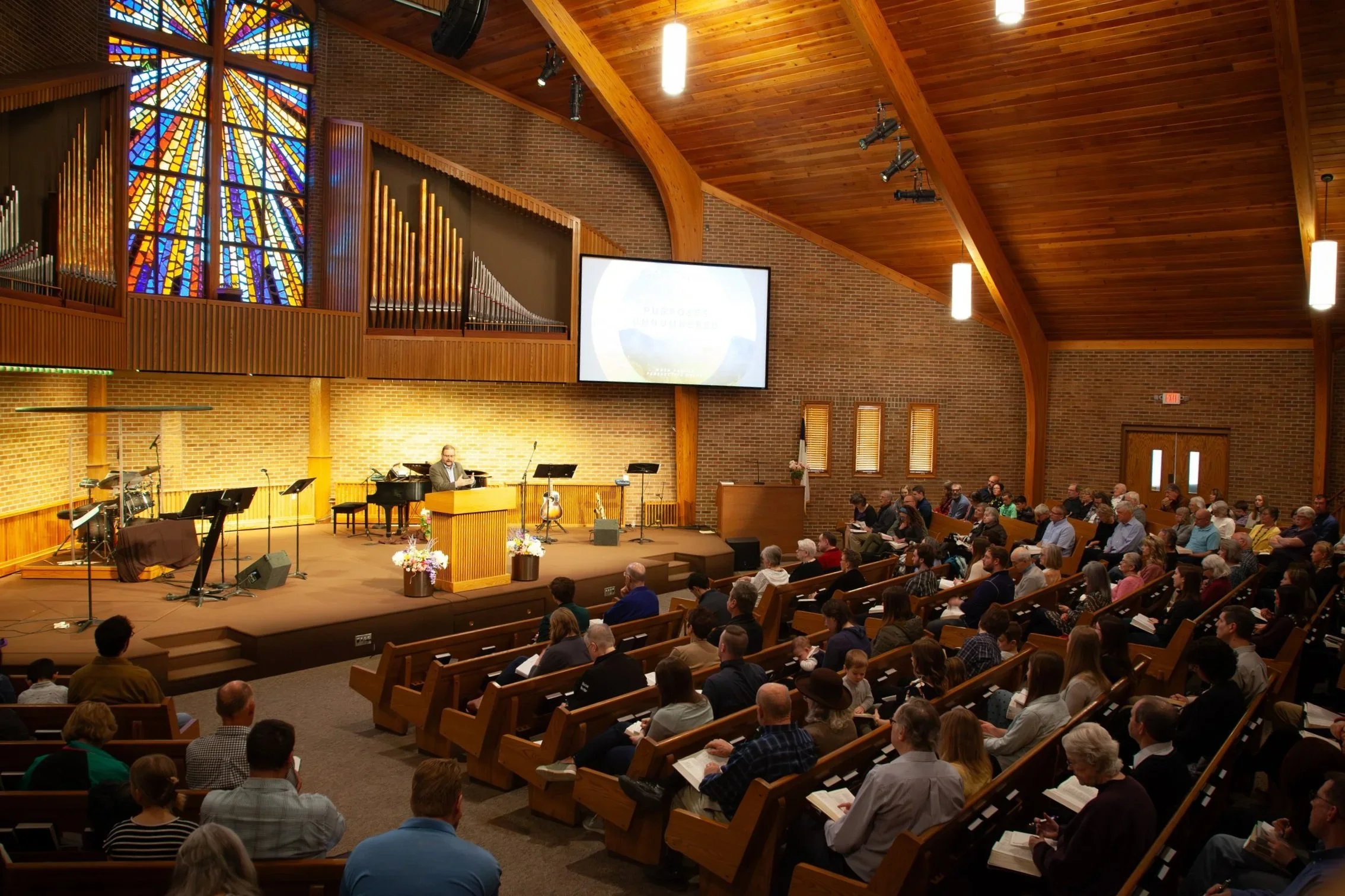 A large congregation gathered inside a church with wooden beams and brick walls for a service or event. The stage at front features musical instruments including a grand piano, drums, and guitars. A pastor or speaker is seated at the piano, while the audience listens attentively. A big stained glass window is visible at the back of the stage, along with hanging lights and a large screen.
