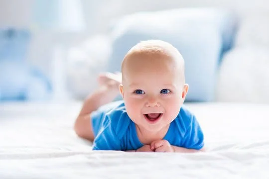 A happy baby lying on its stomach on a bed, wearing a blue shirt.
