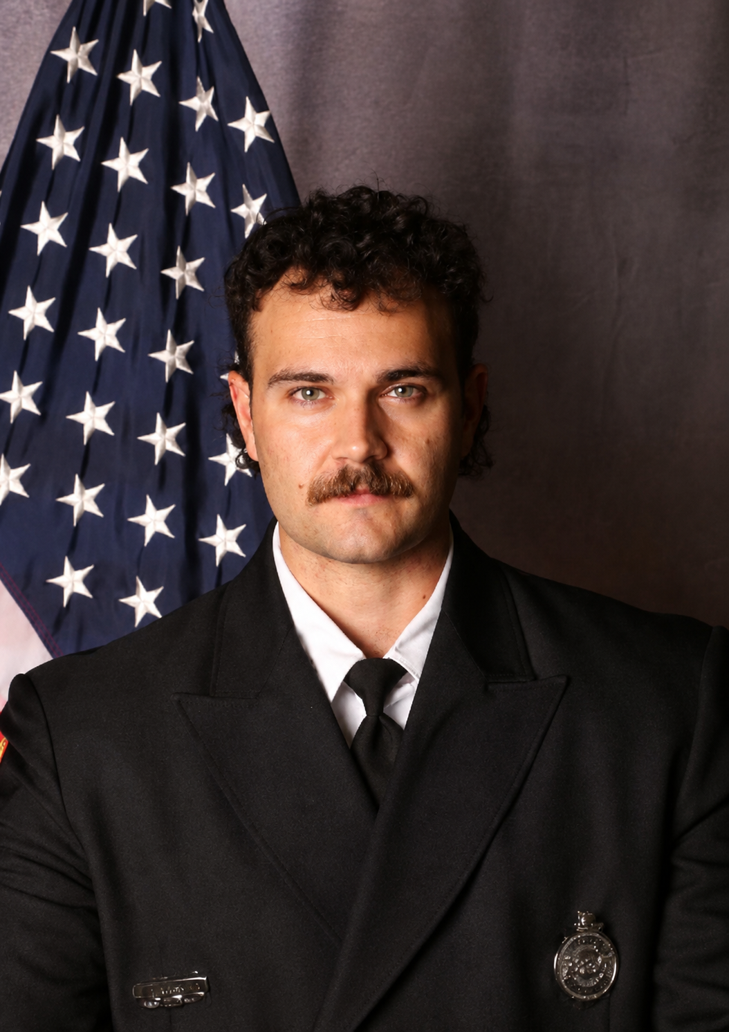 A police officer with a mustache and curly hair, wearing a black uniform with a badge, standing in front of an American flag.