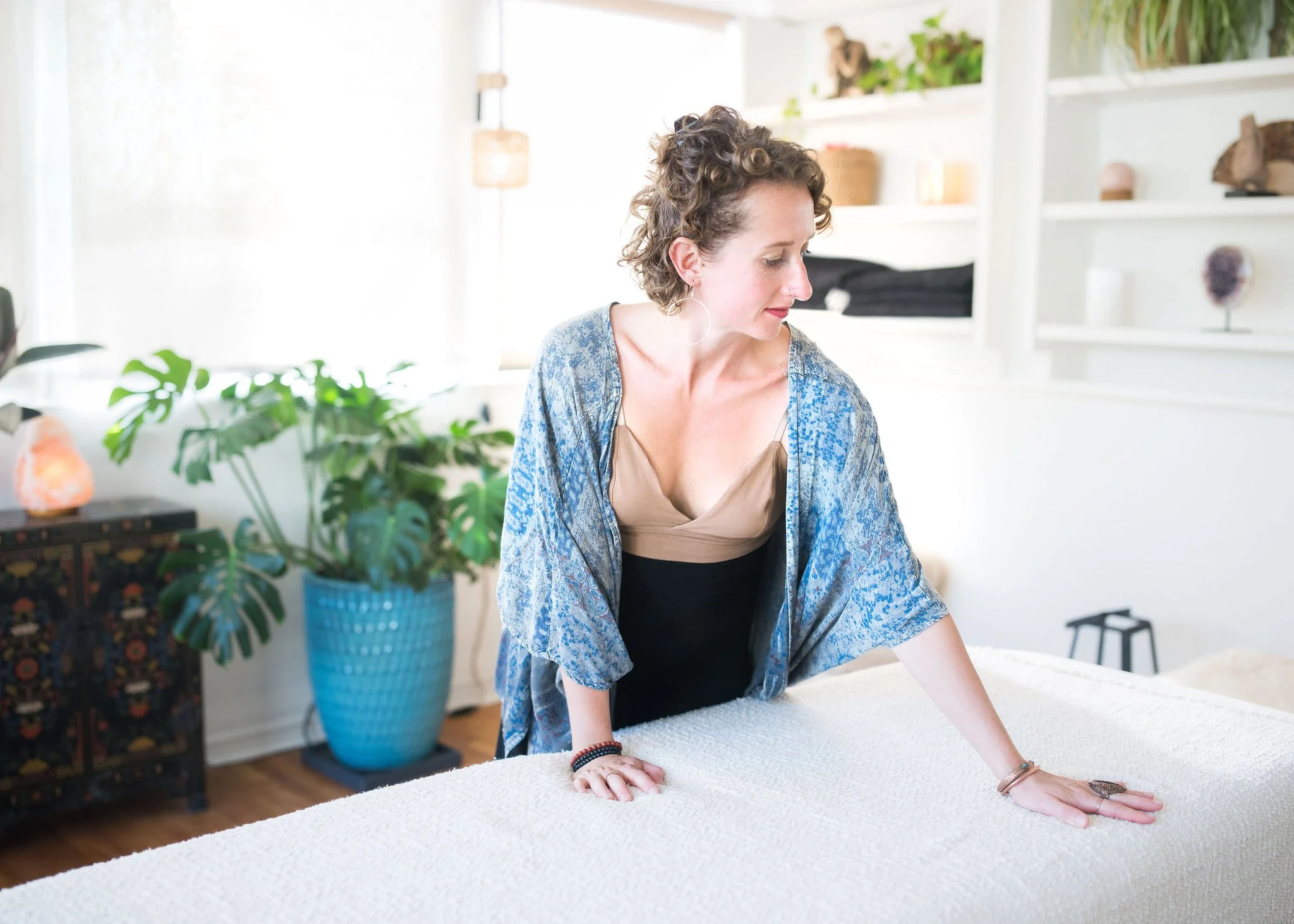Woman with curly hair and hoop earrings leaning on a bed in a bright bedroom with plants and shelves.