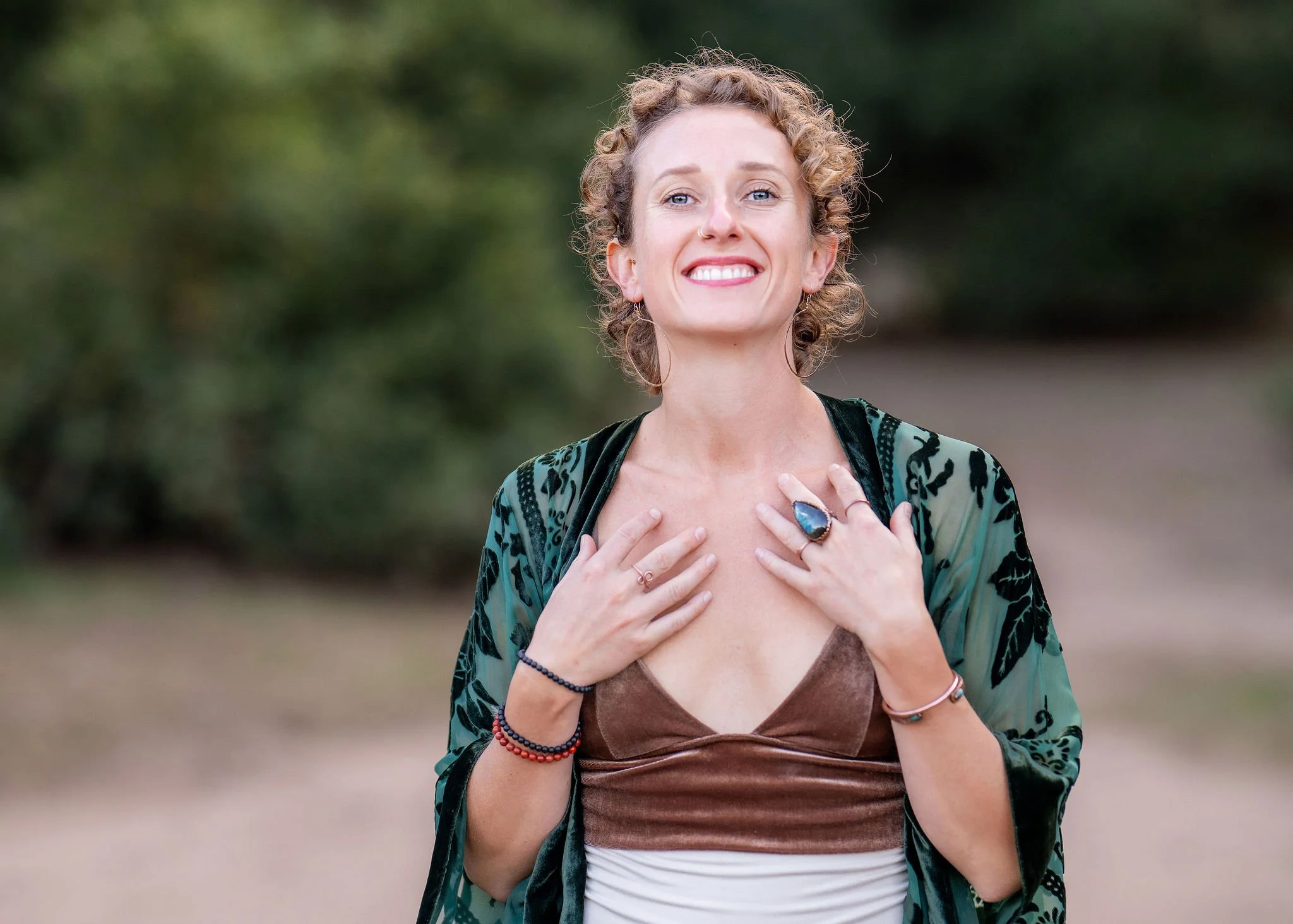 A woman with curly hair and blue eyes smiling, holding her chest with both hands, wearing a patterned green jacket over a brown top, standing outdoors with a blurred green background.
