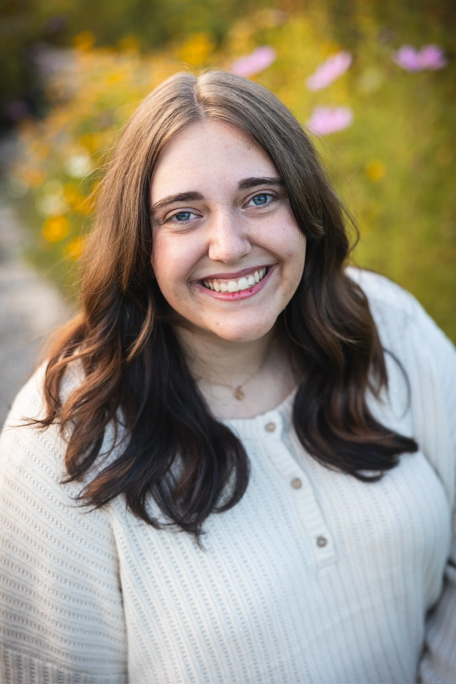 A young woman with long wavy brown hair and blue eyes, smiling, wearing a white sweater with buttons, outdoors during fall with blurred colorful foliage in the background.
