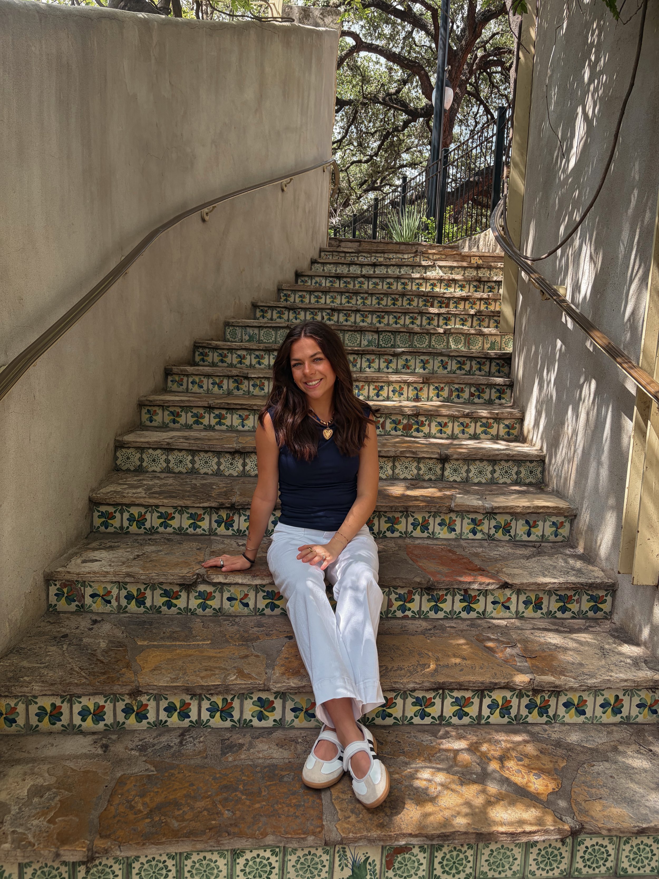 A woman sitting on decorative tiled steps outdoors, smiling, with large trees in the background.