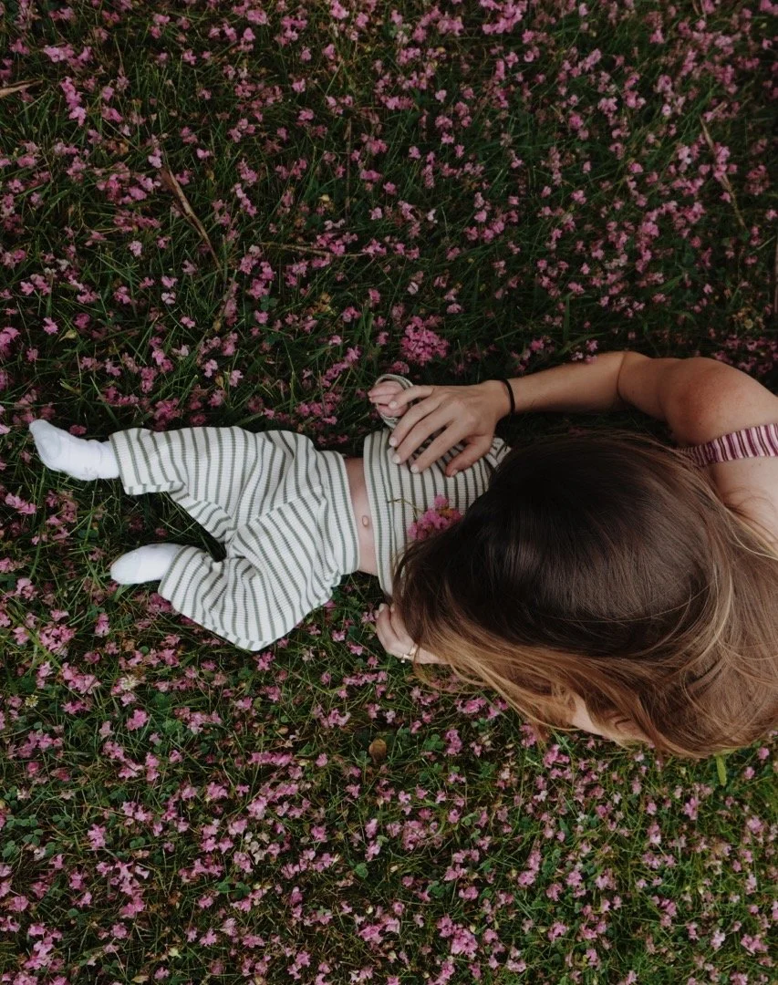 A woman lying on her side in a field of pink flowers, resting her head on her hand, with a small child lying next to her, dressed in white socks, striped pants, and a striped top.