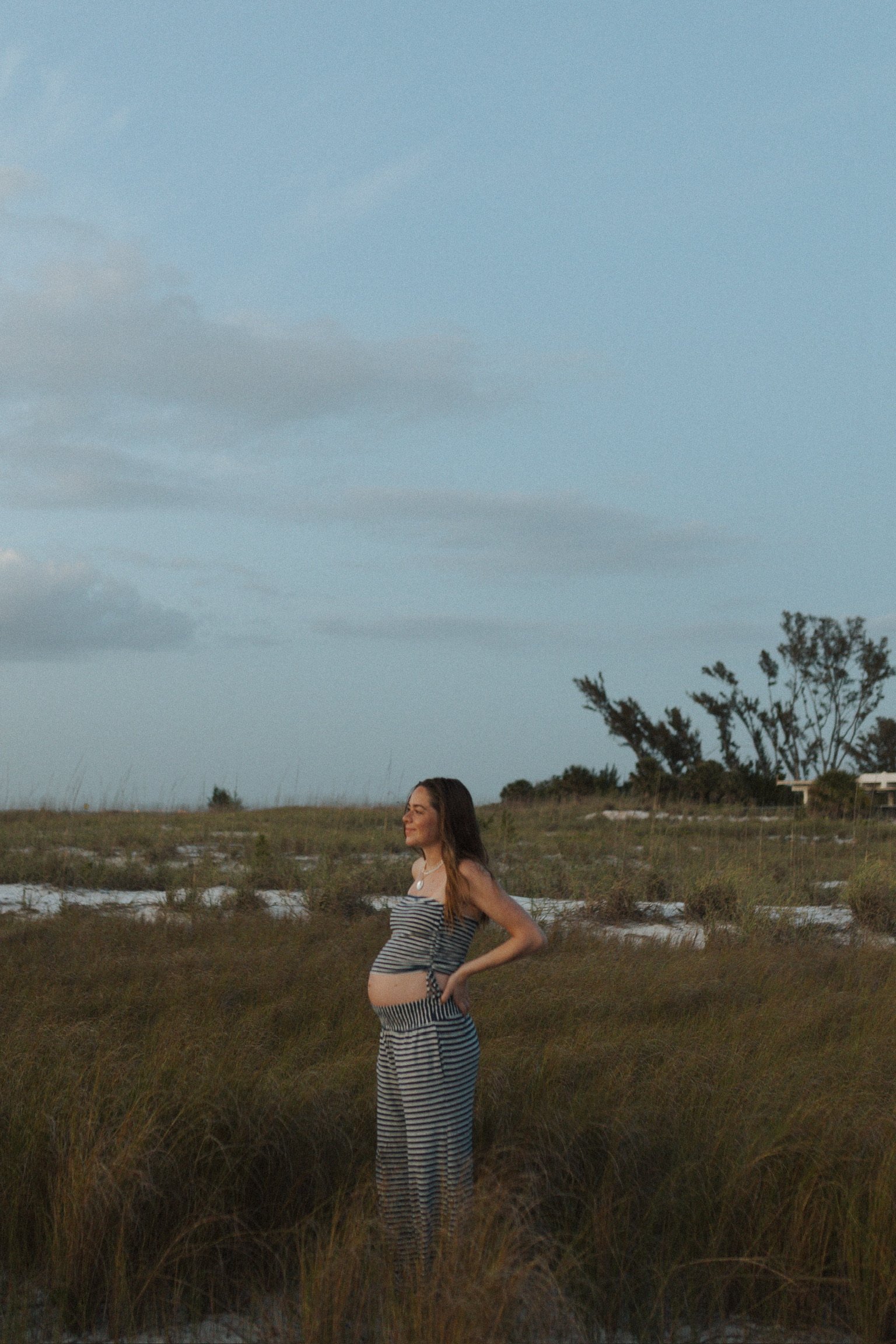A pregnant woman with long brown hair standing in a grassy field during sunset, wearing a striped dress with bare shoulders, smiling and with her hands on her hips, with trees and buildings in the distance under a blue sky with some clouds.