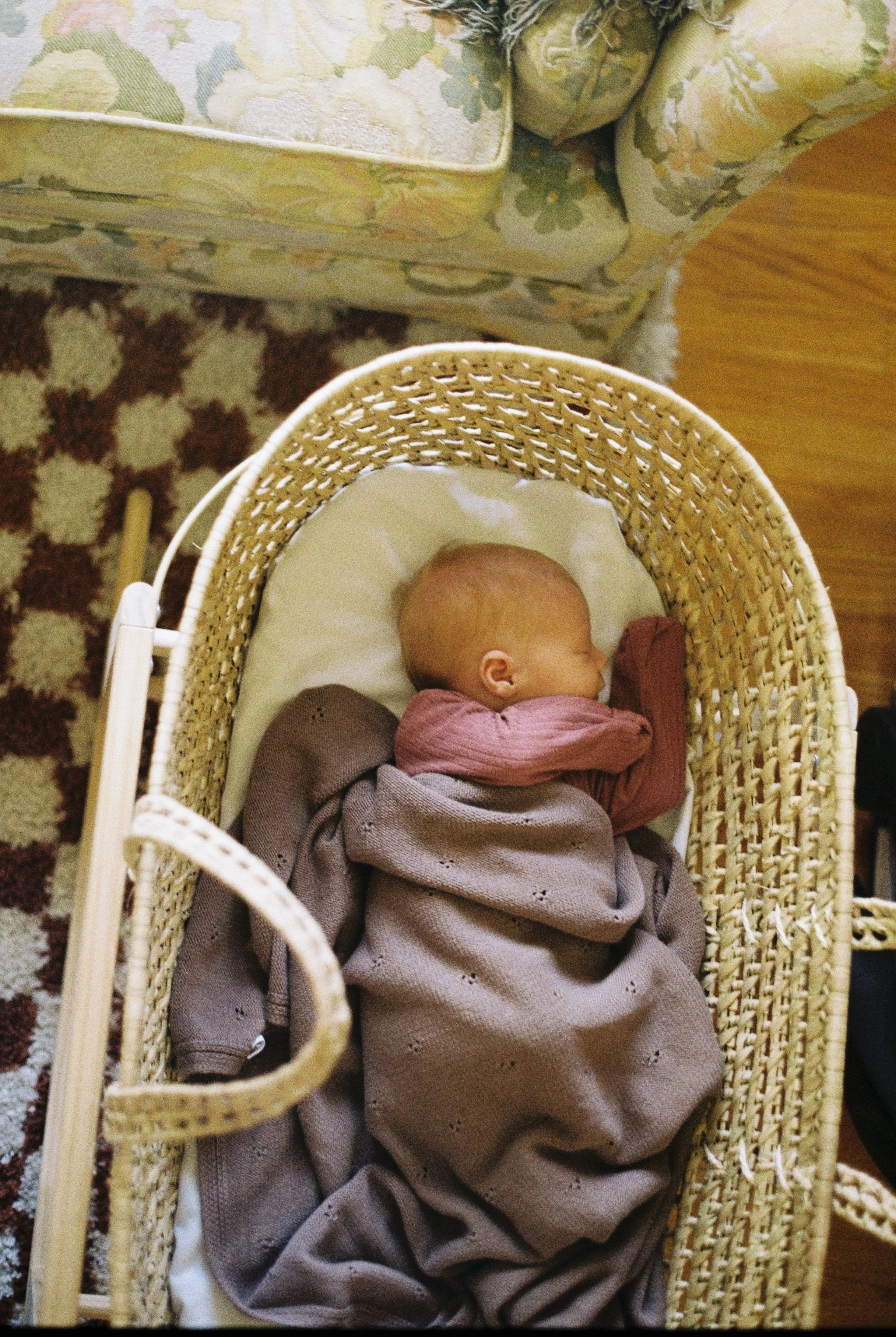 A sleeping baby lying in a woven bassinet with a white mattress, wearing a pink top and gray pants, in a room with a patterned area rug and a floral upholstered armchair.