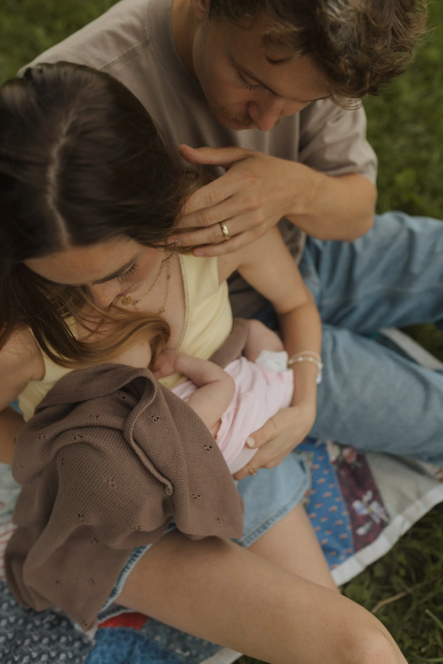 A man, woman, and child sitting outdoors on a blanket in a grassy area. The man is leaning over the woman and child, looking at the child with a caring expression. The woman is holding the child and their faces are close together, creating an intimate family moment.