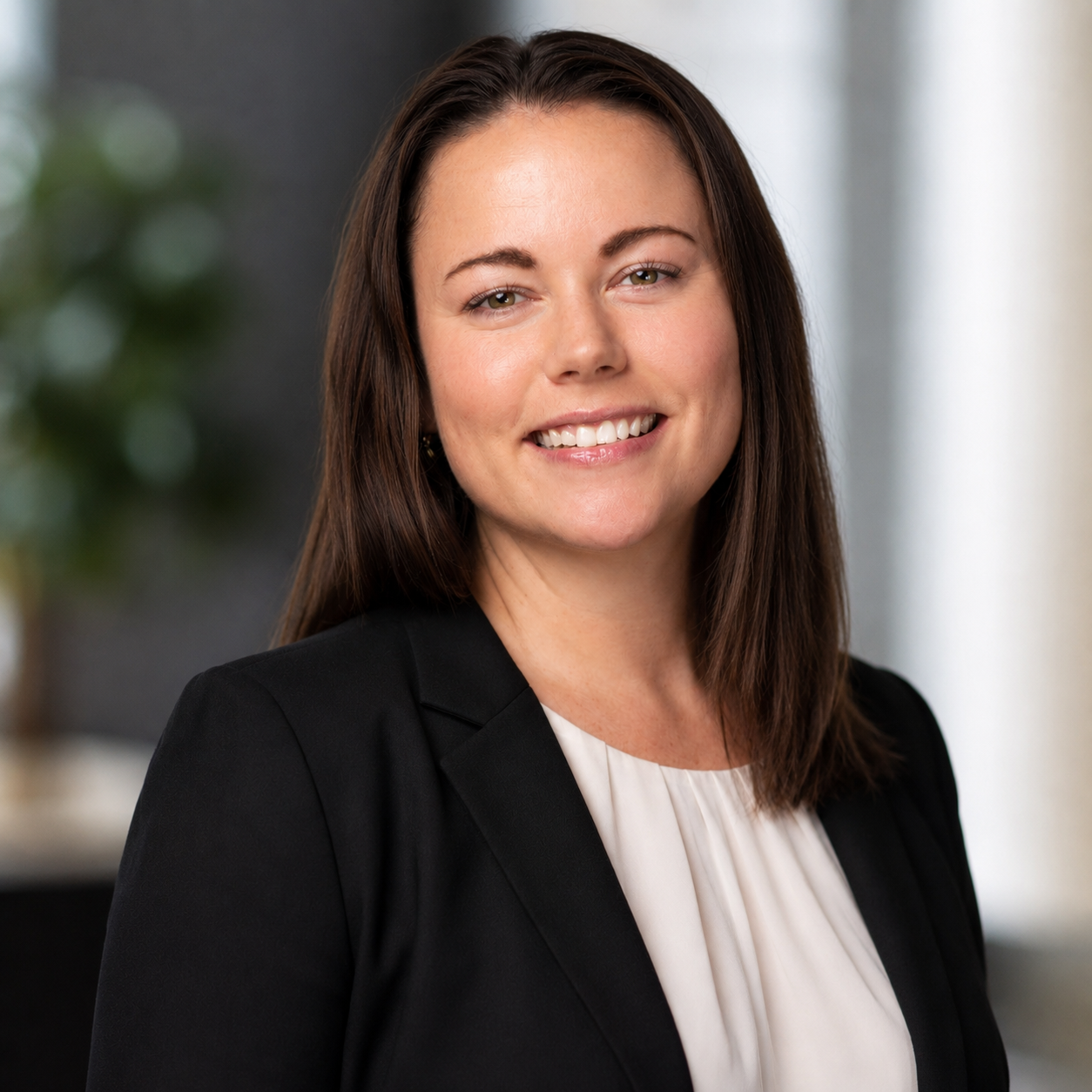 A professional woman with shoulder-length brown hair smiling, wearing a black blazer over a white blouse, in an office setting.