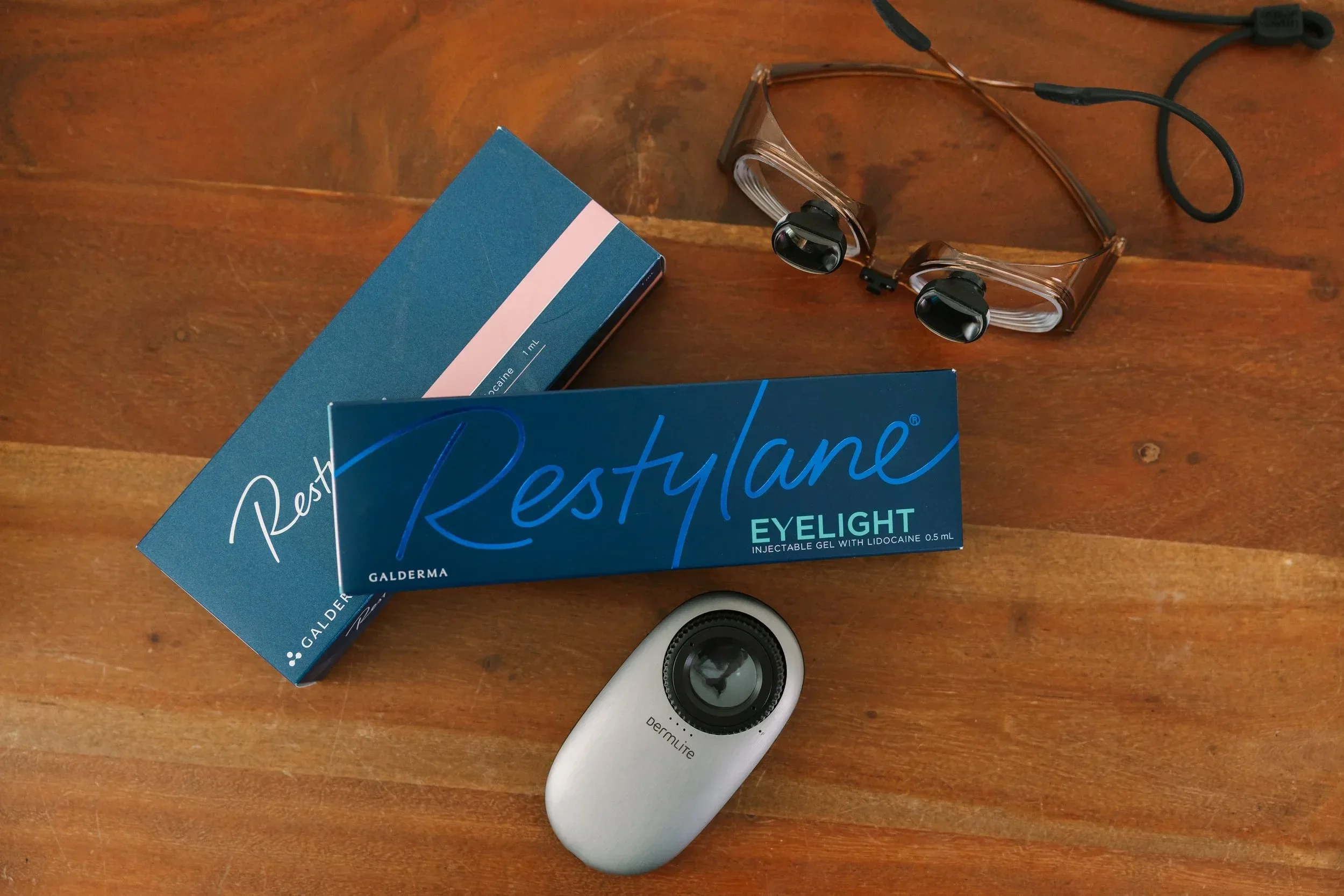 Close-up of a wooden table with a box of Restylane Eyelight, a pair of transparent glasses with black earpieces, and a white Dermatim device.