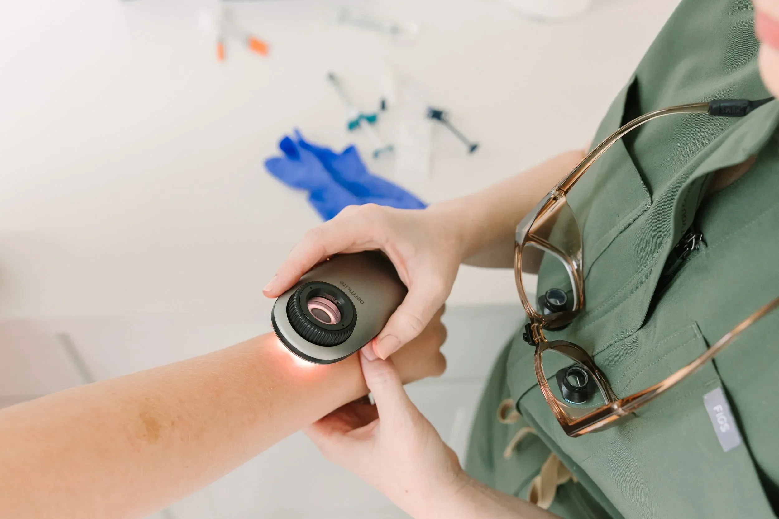 A healthcare professional wearing green scrubs and glasses, using a handheld device that emits a red light on a patient's wrist, with a blurred background including medical tools and a stethoscope.