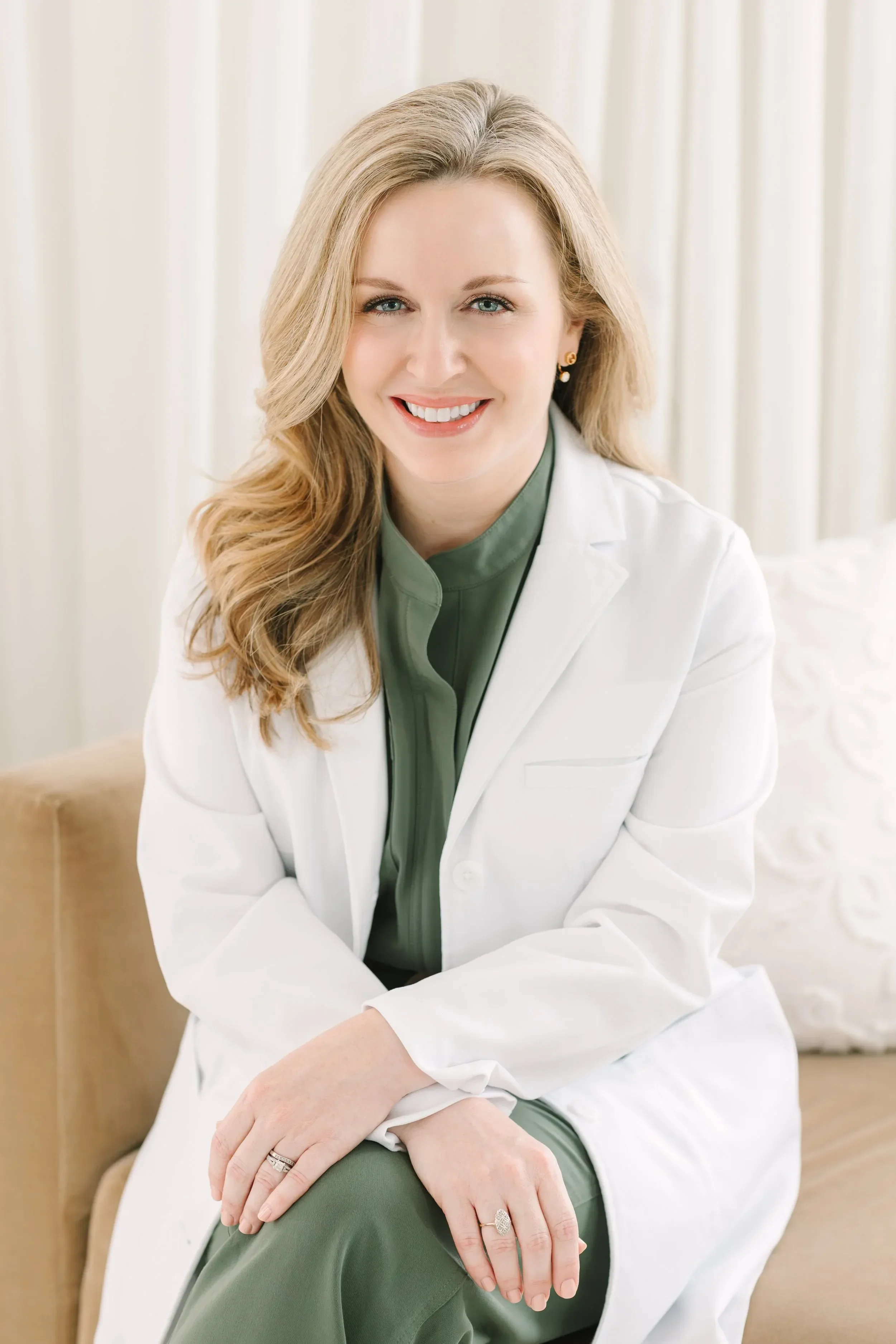 Dr. Lesley Starnes with blonde hair, wearing a white blazer and green top, sitting on a beige sofa in front of beige curtains, smiling at the camera, with rings on her fingers and earrings.