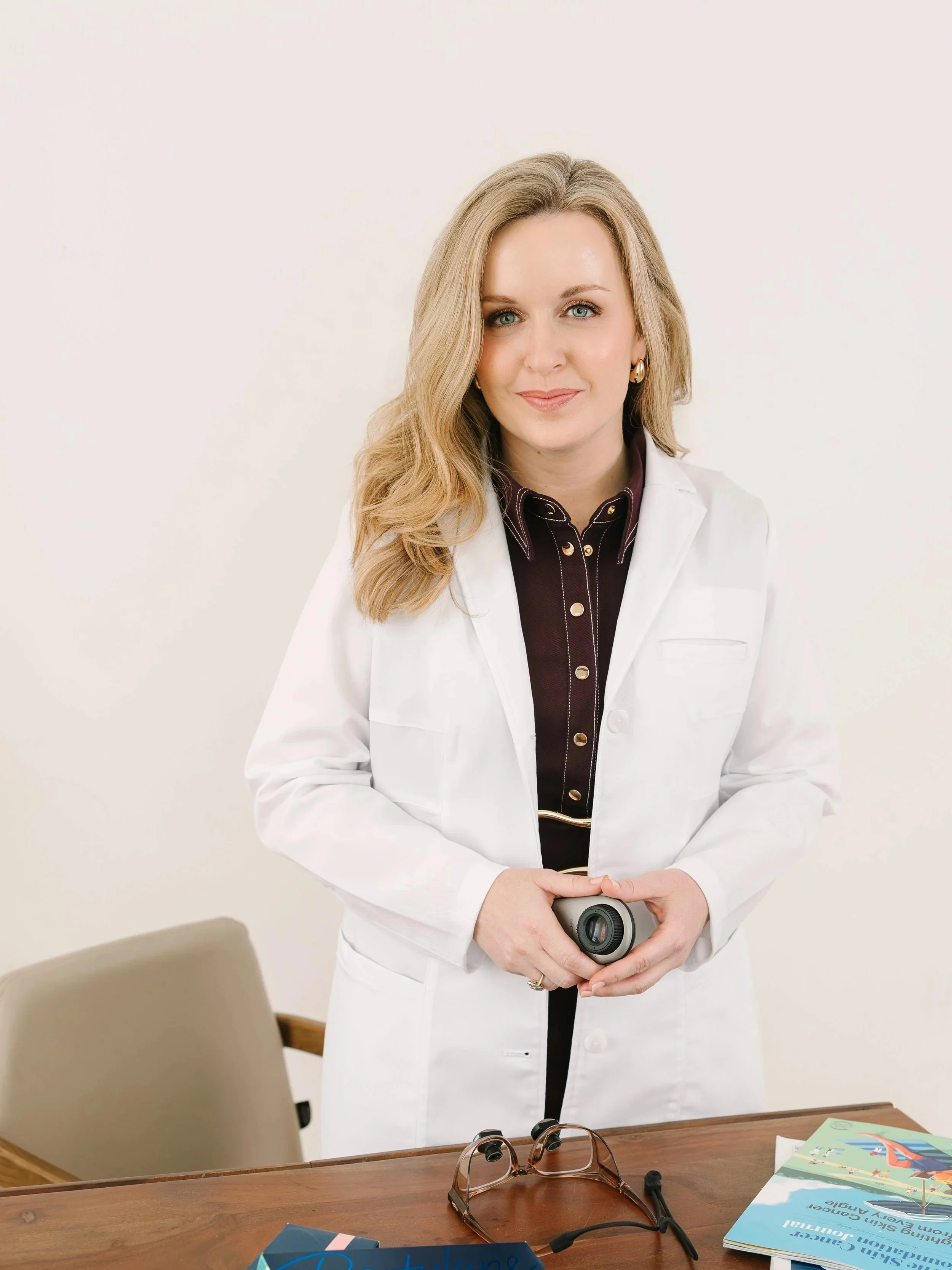 Dr. Lesley Starnes with blonde hair and blue eyes in a white doctor's coat holding a camera, standing in a room with a wooden table. On the table, there are sunglasses, a magazine, and some papers.