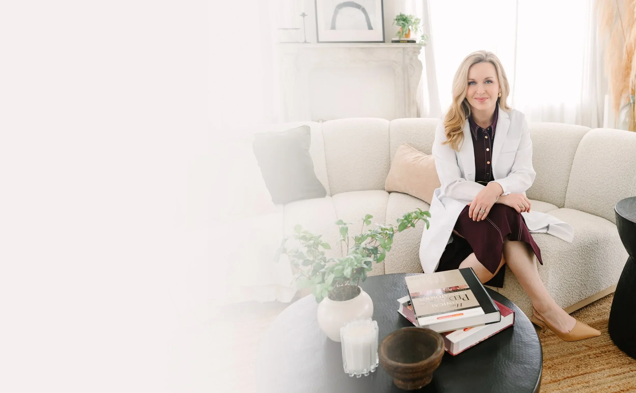Dr. Lesley Starnes in a white blazer sitting on a cream sofa in a bright living room with books, plants, and artwork, smiling at the camera.