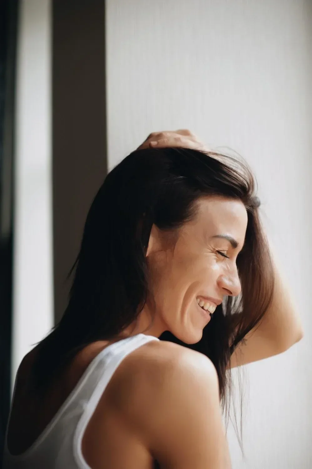 A woman with dark hair smiling and laughing with her eyes closed, wearing a white tank top, with her right hand resting on her head.