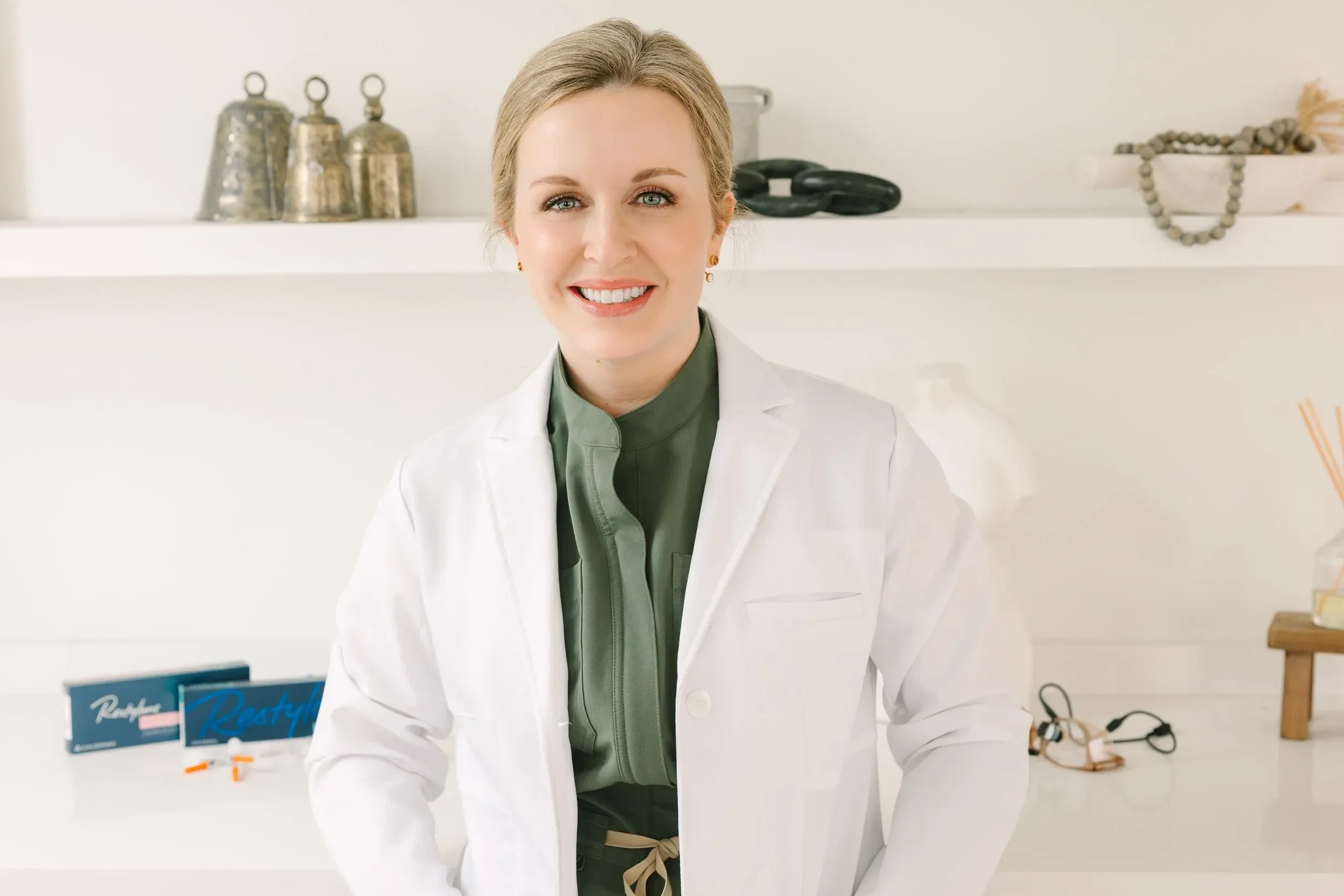 Dr. Lesley Starnes smiling in a white coat standing in front of a shelf with decorative items and health products.