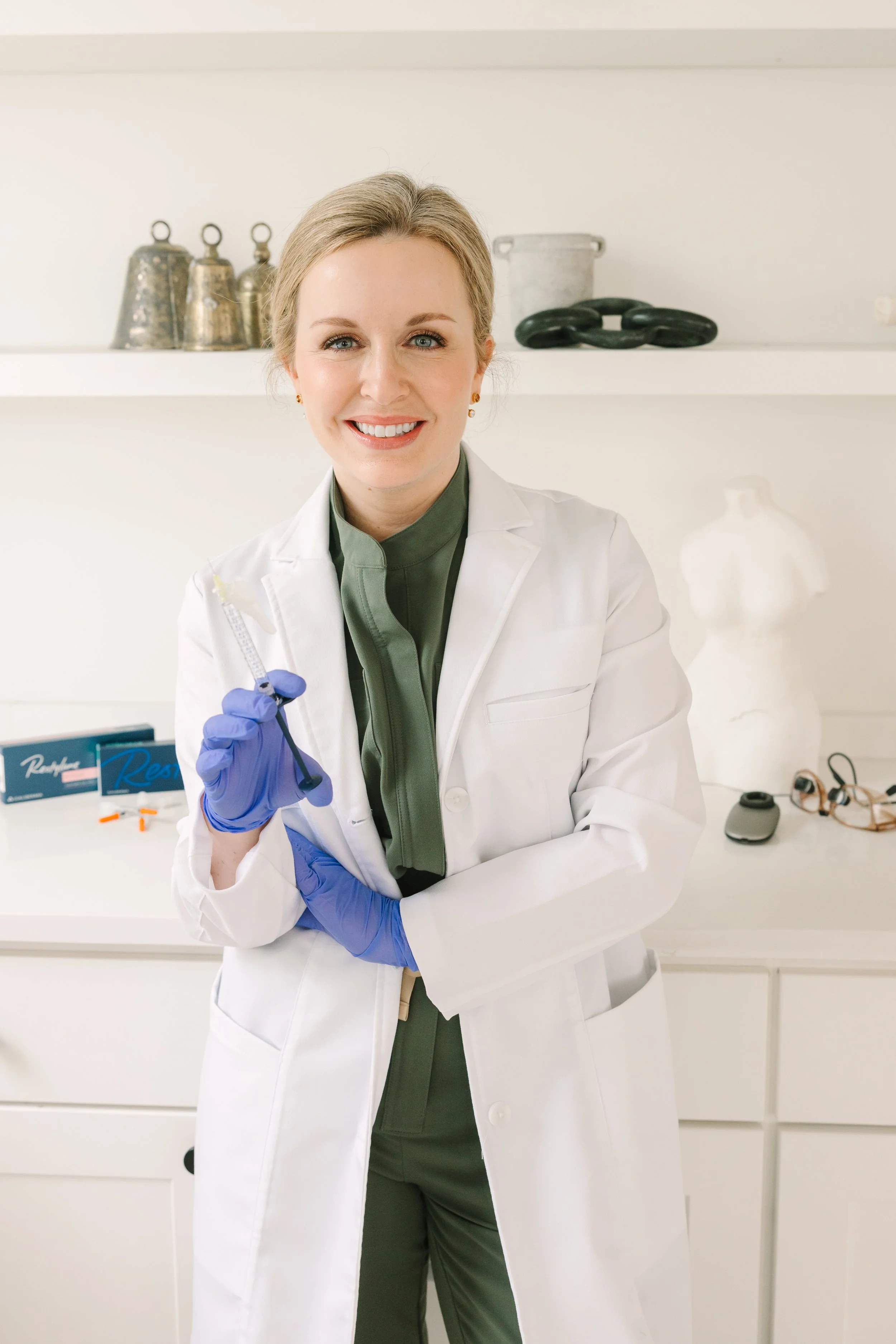 Dr. Lesley Starnes smiling in a white coat and green scrubs holding a syringe, standing in her clinic.