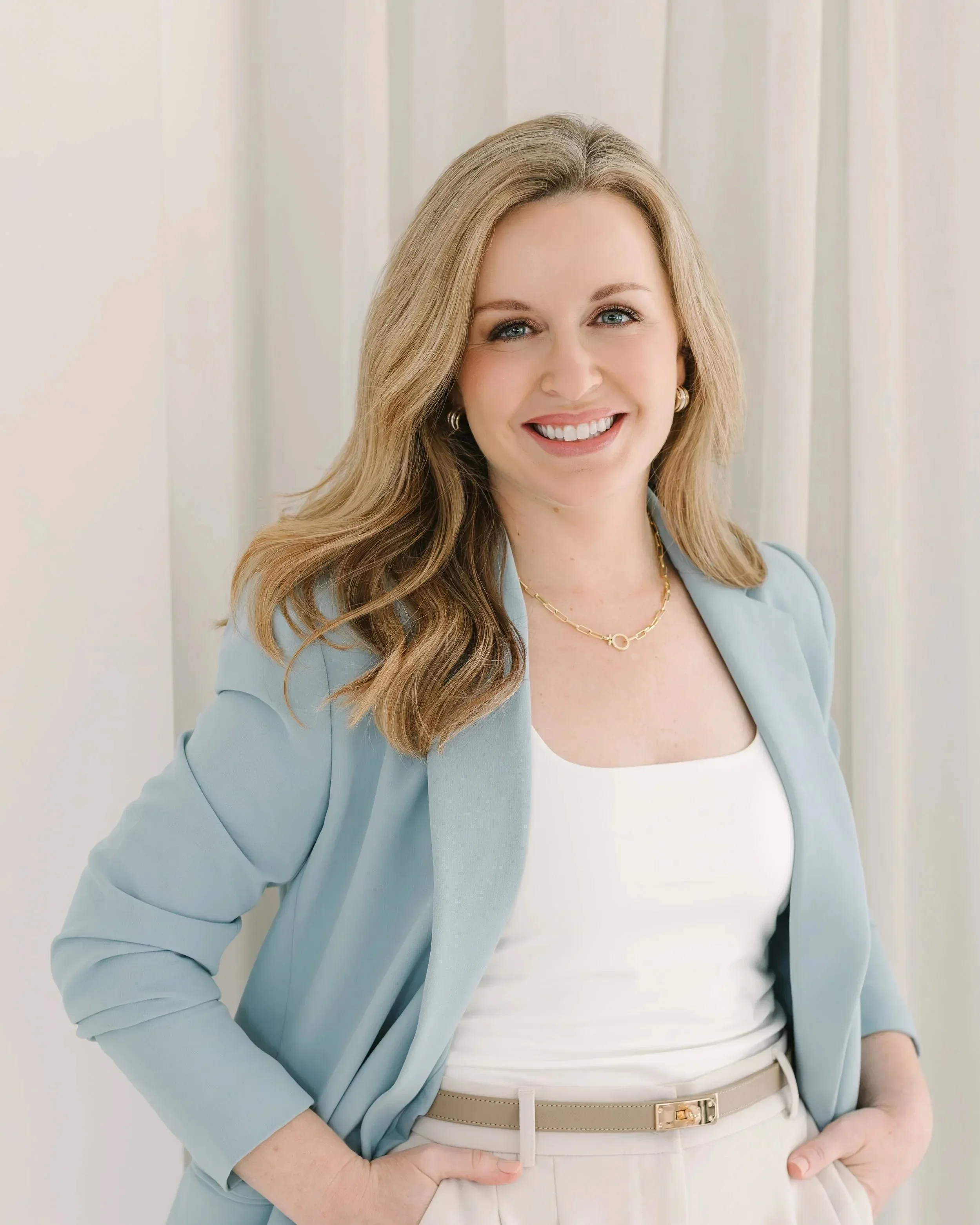 Close-up of Dr. Lesley Starnes wearing a light blue blazer, white top, and beige belt, smiling with hands in her pockets, standing in front of a neutral background.