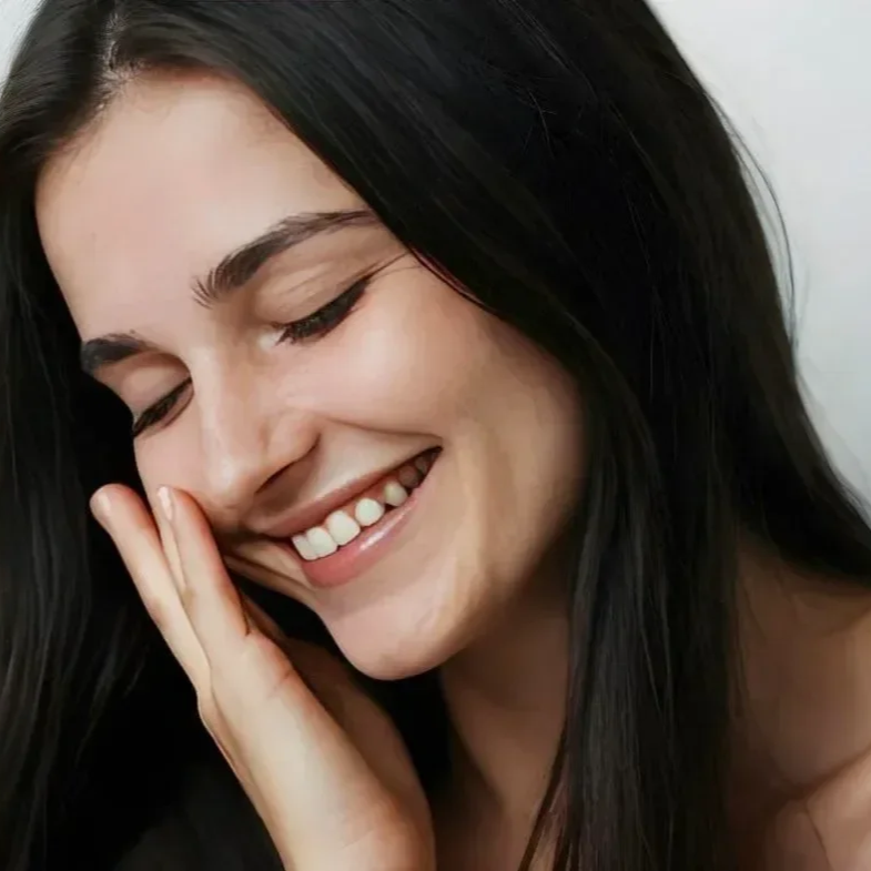 Close-up of a smiling woman with long black hair, gently touching her cheek with her hand, eyes closed.