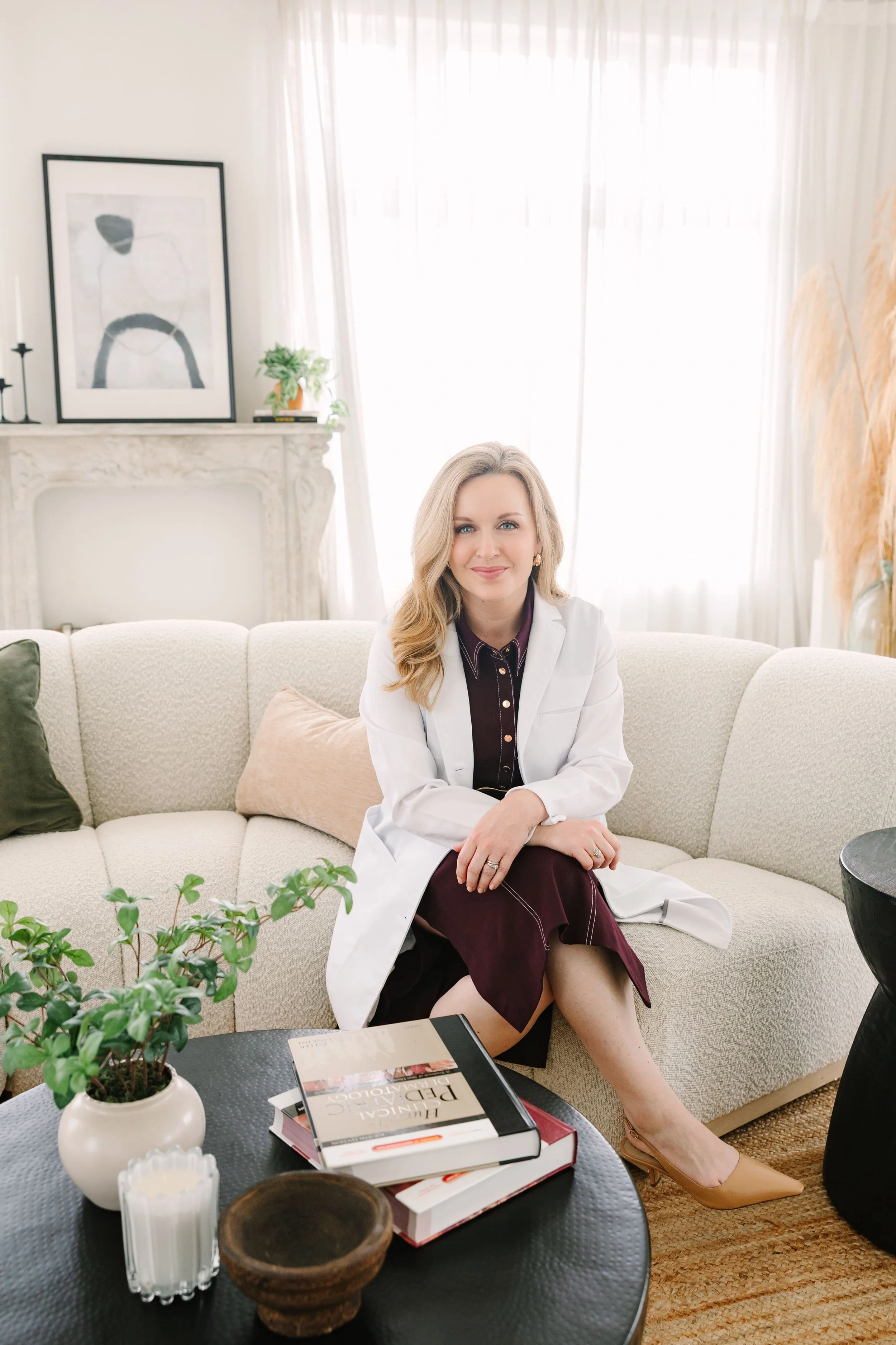 Dr. Lesley Starnes in a white coat sitting on a beige sofa in a bright living room, with books, plants, and decorative items around her.