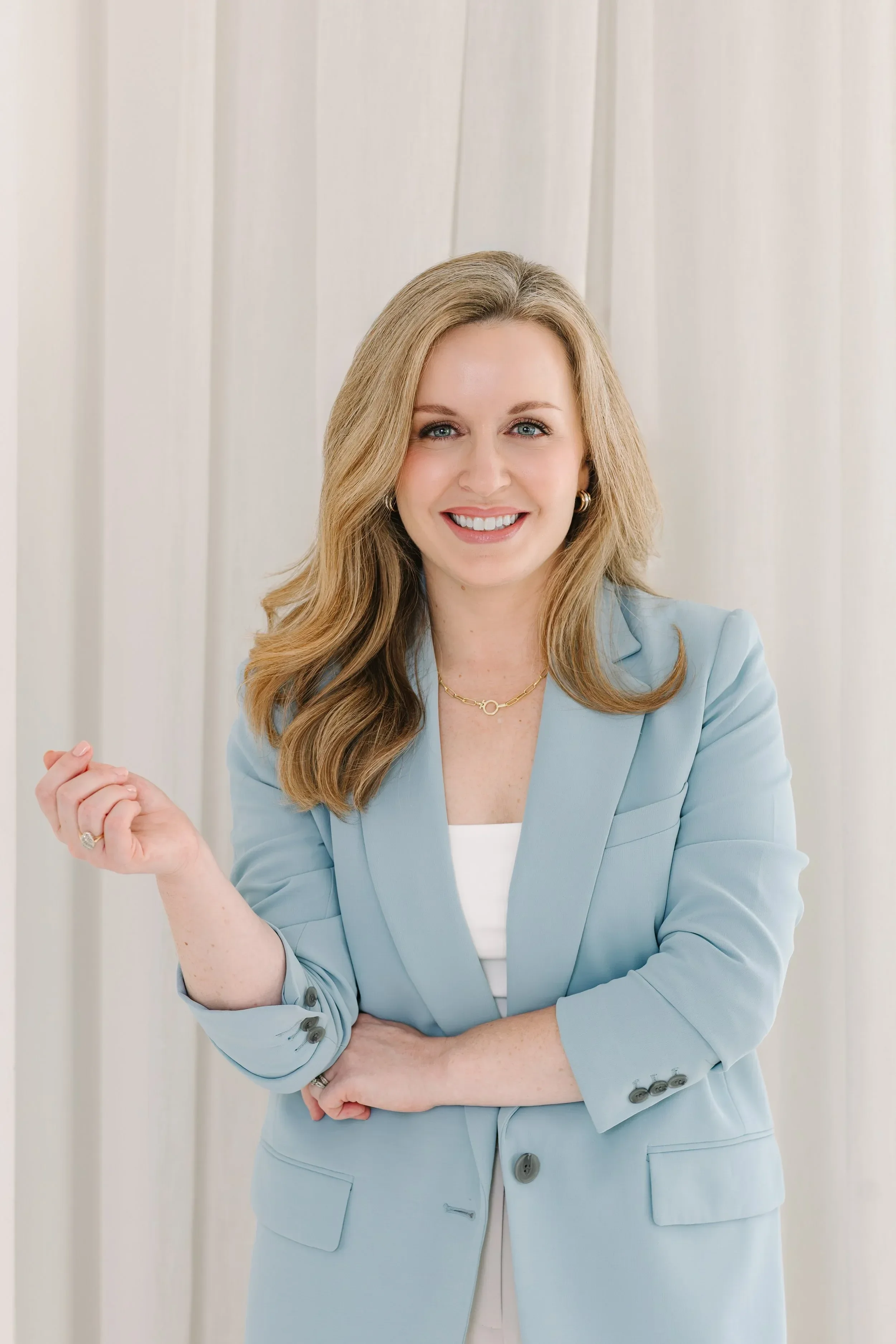 Dr. Lesley Starnes with long light brown hair, wearing a light blue blazer and a white top, smiling at the camera with a background of white vertical blinds.