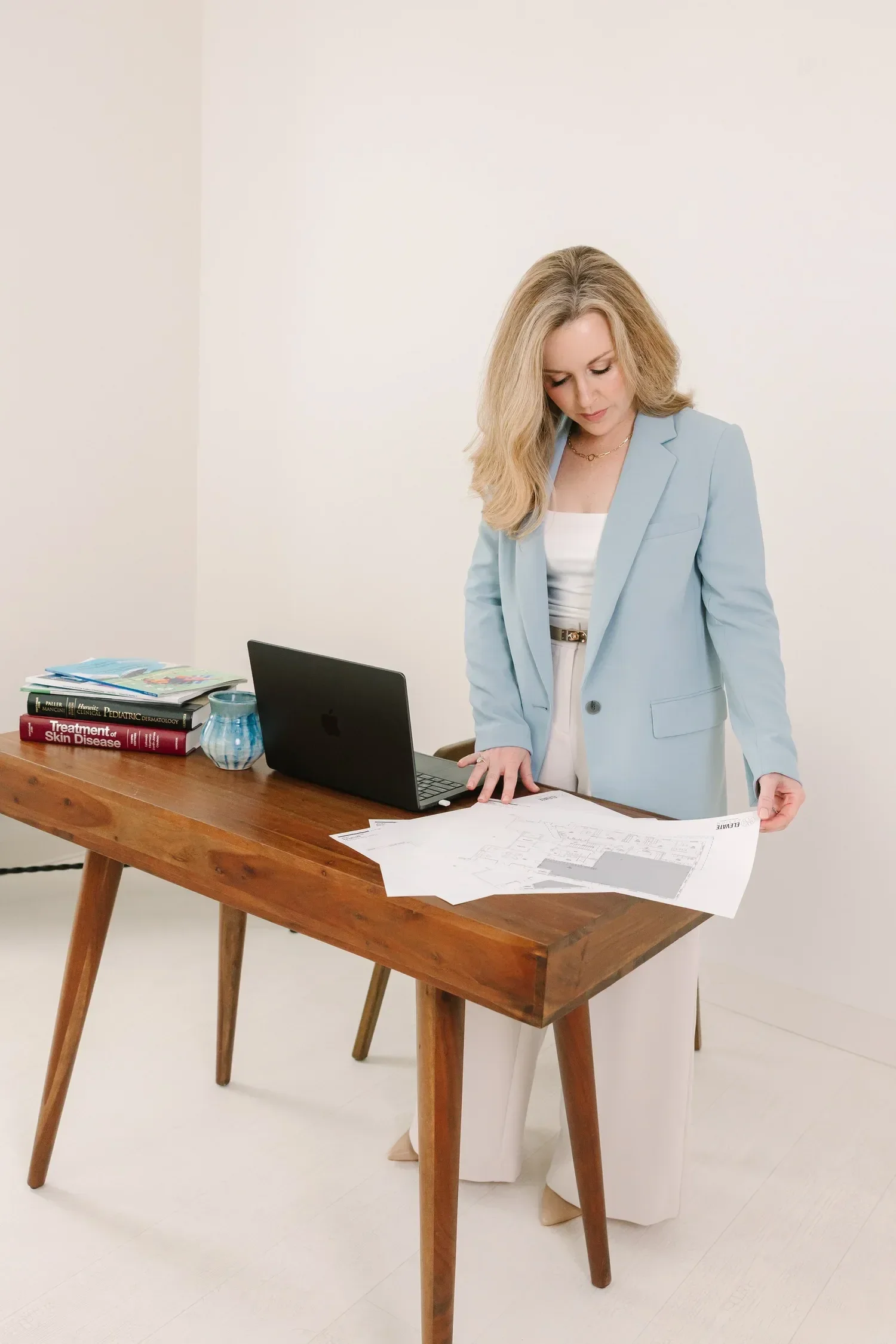 Dr. Lesley Starnes in a light blue blazer and cream pants stands at a wooden table, examining assessment papers. The table holds a laptop, a stack of books, a ceramic mug, and some papers. The background is plain and white.