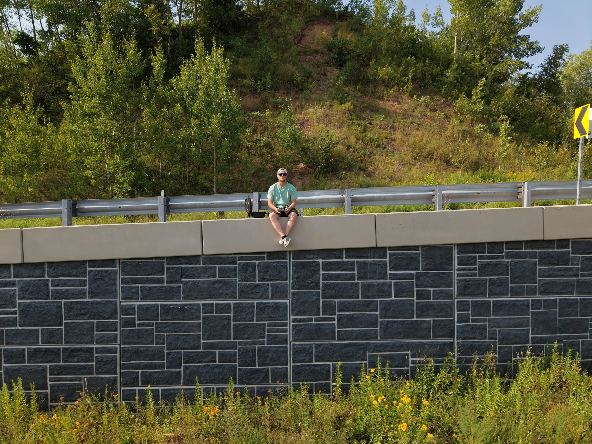 A man wearing sunglasses and a teal shirt sitting on a concrete barrier beside a highway, with green trees and a hillside in the background, and a yellow road sign indicating a curve.