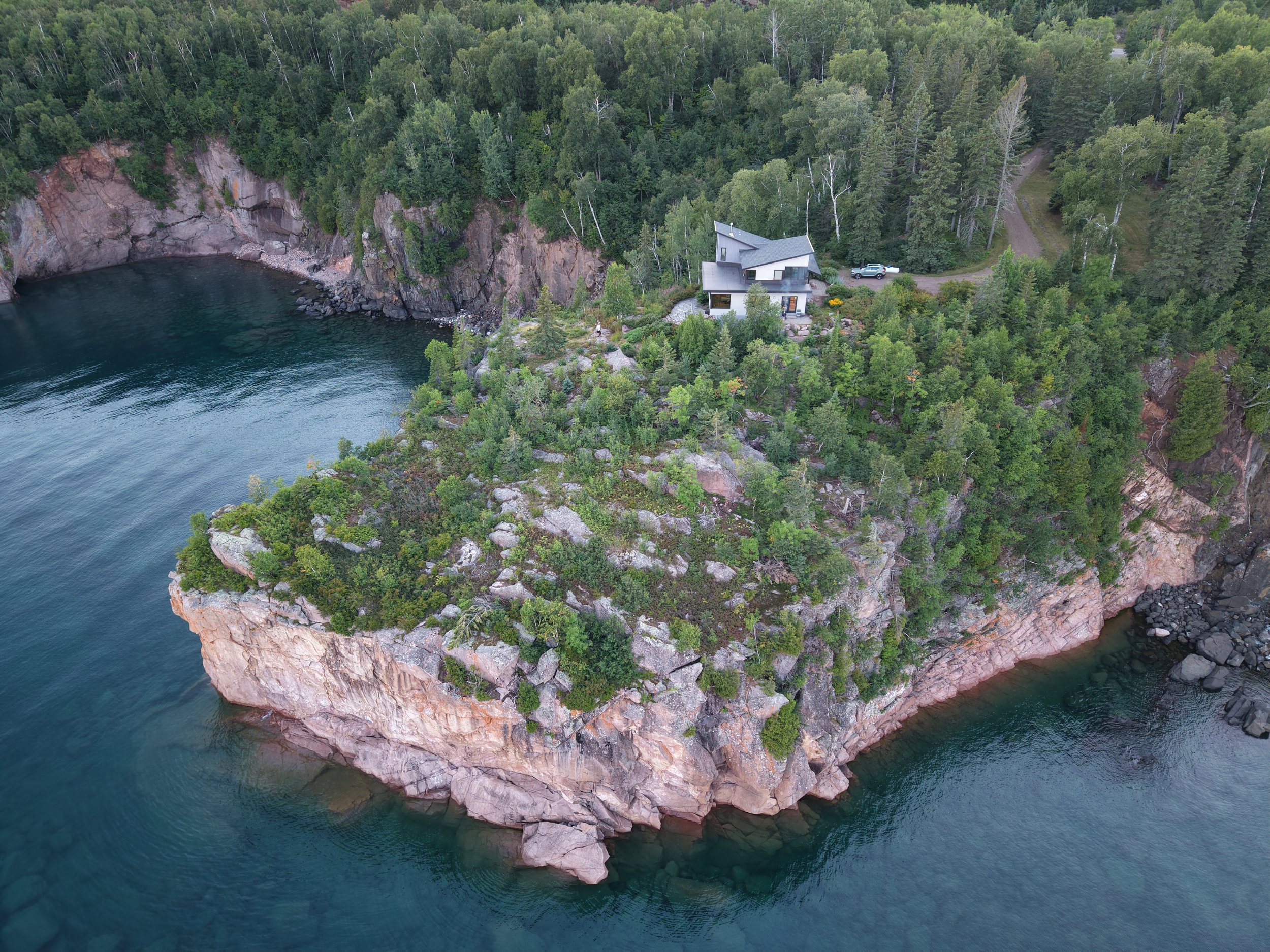 Aerial view of a house surrounded by dense forest on a rocky peninsula extending into a body of water.