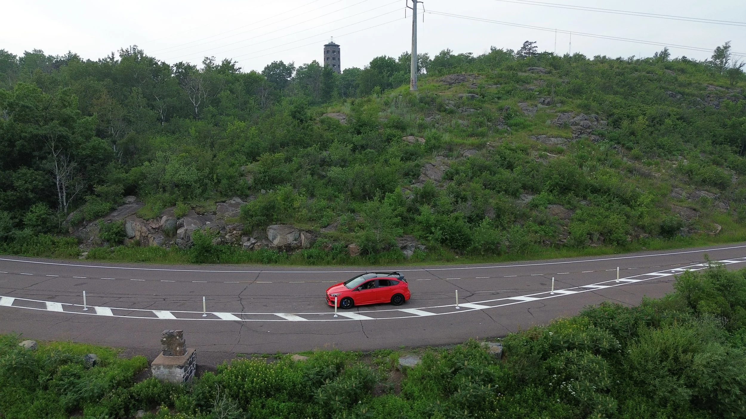 A red car parked on the side of a curved road with white dashed lines and barriers, surrounded by green trees and rocks, with a tall structure and power lines in the background.