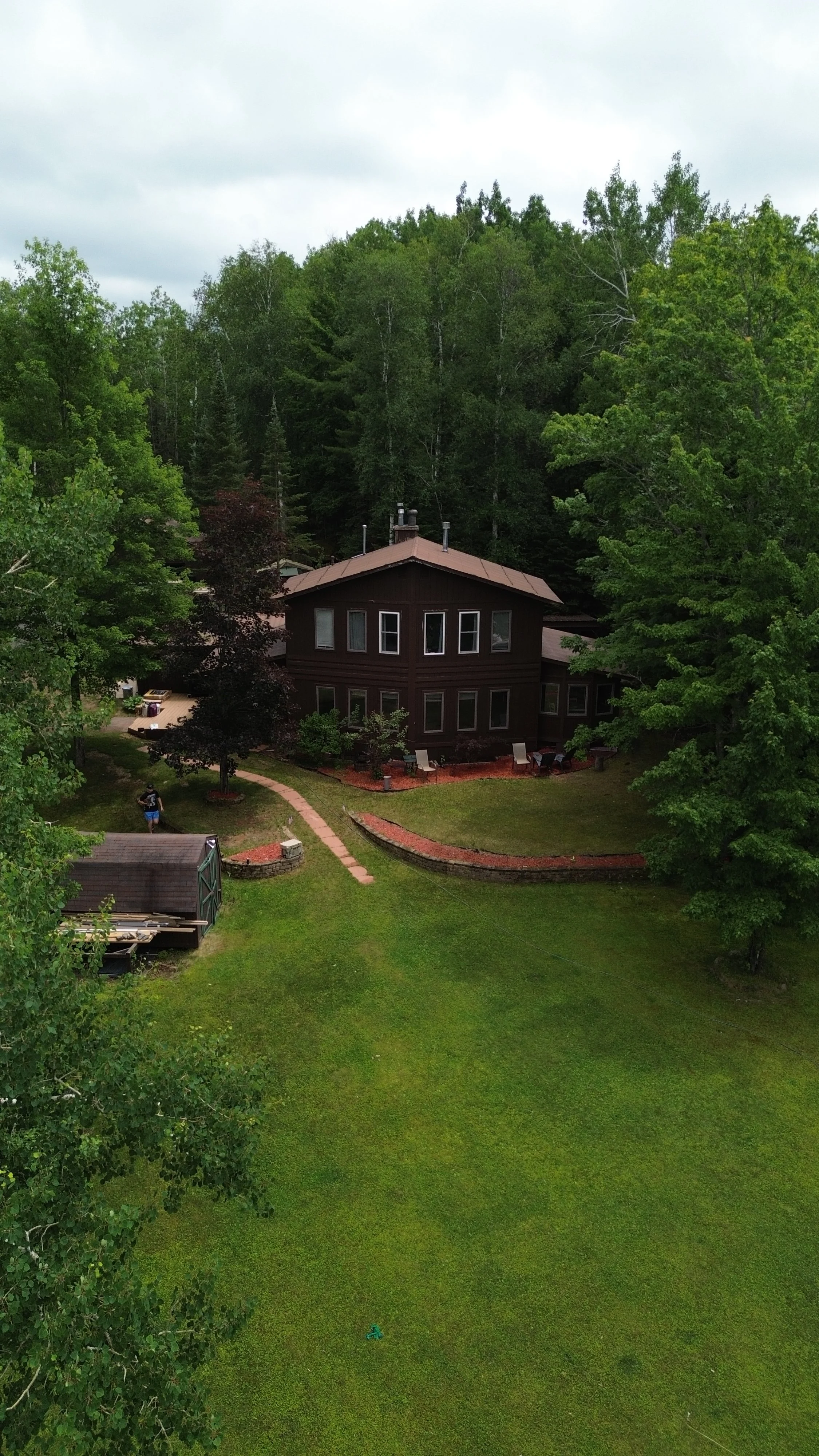 A two-story house with dark brown exterior in a green backyard surrounded by trees, with a paved walkway, lawn chairs, and a person walking along the path.