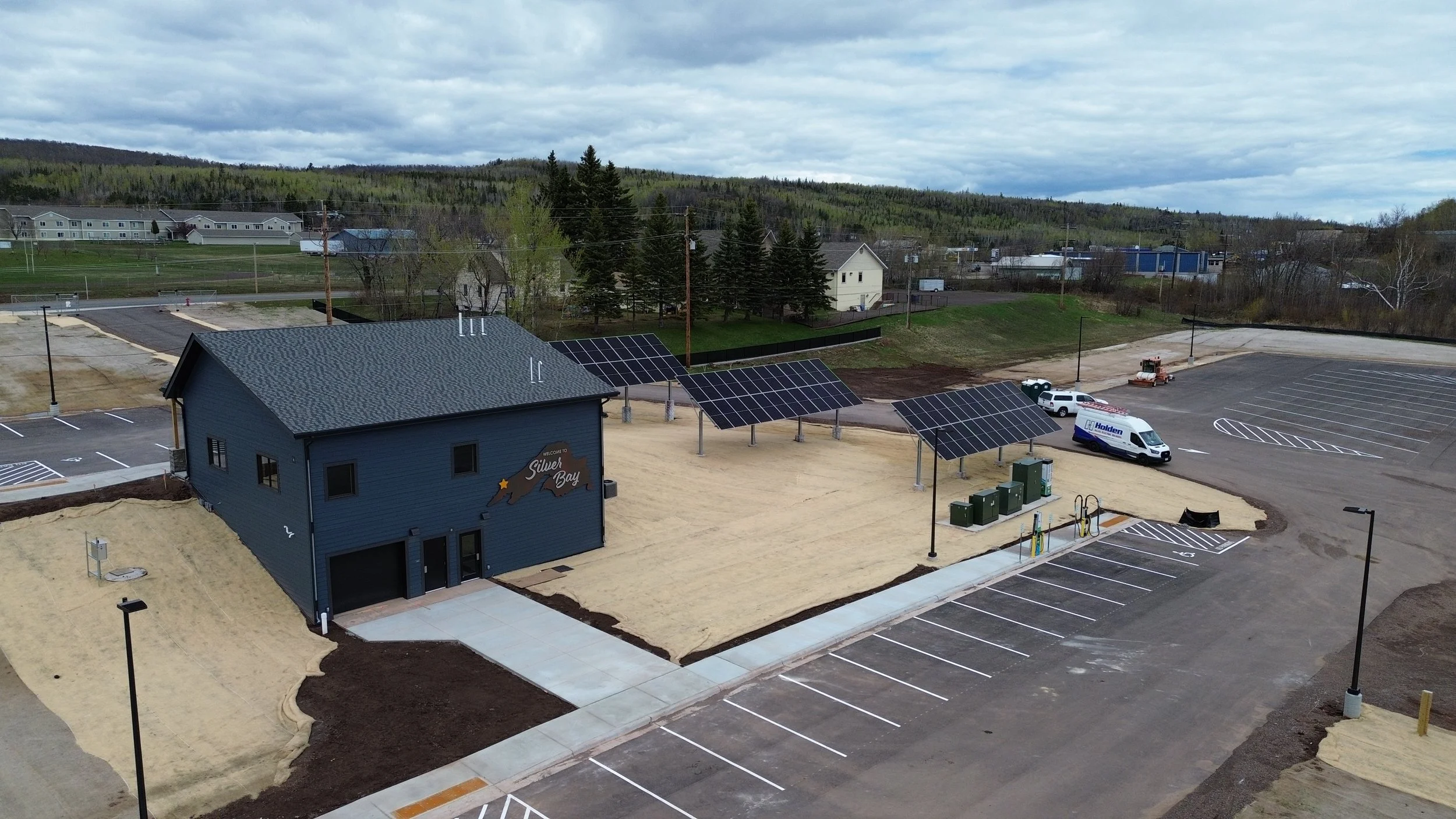 An area under construction with a building labeled 'Silver Bay,' solar panels, and a parking lot with some parked vehicles, including a maintenance vehicle. Hills and houses are visible in the background under a cloudy sky.