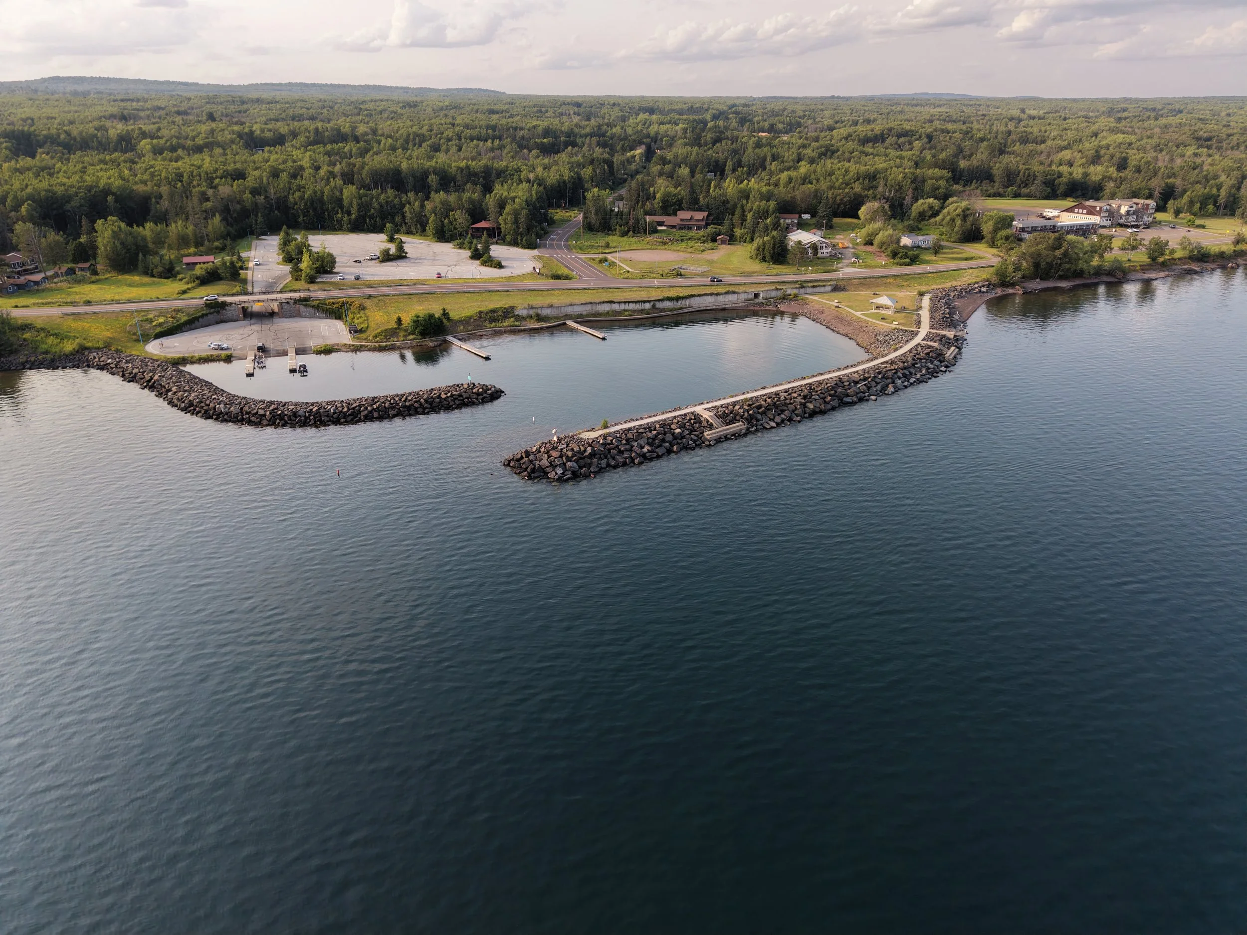 Aerial view of a coastal area with a small marina, parking lot, roads, and greenery, overlooking a large body of water.
