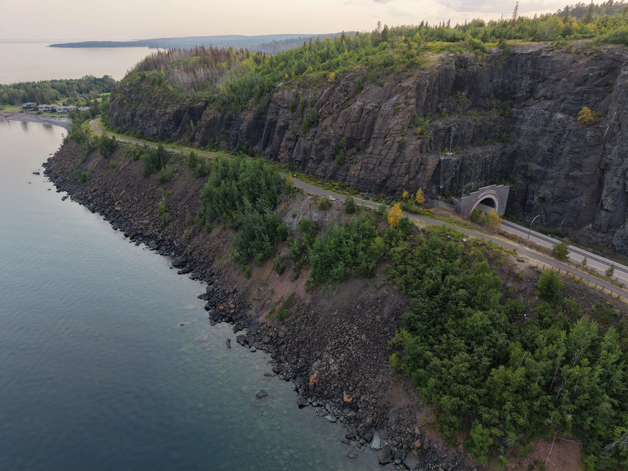 Aerial view of a cliffside with a road, trees, and a tunnel entrance. Water body at the base of the cliff with rocks along the shoreline.