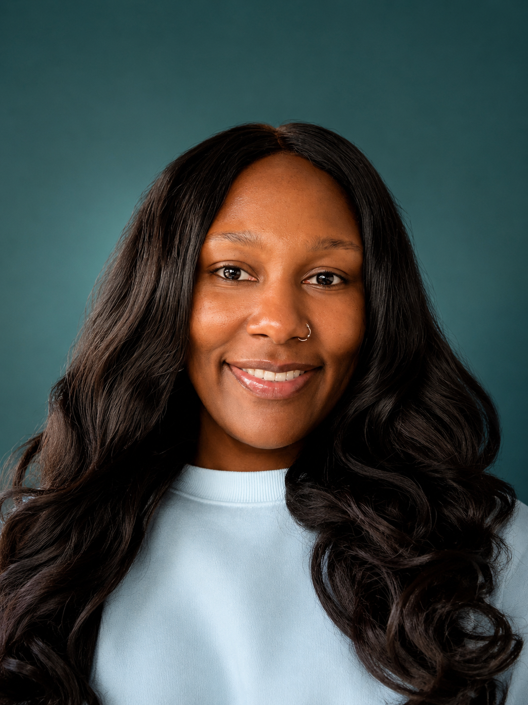 Close-up portrait of a smiling Black woman with long black curly hair, wearing a light blue shirt, against a teal background.