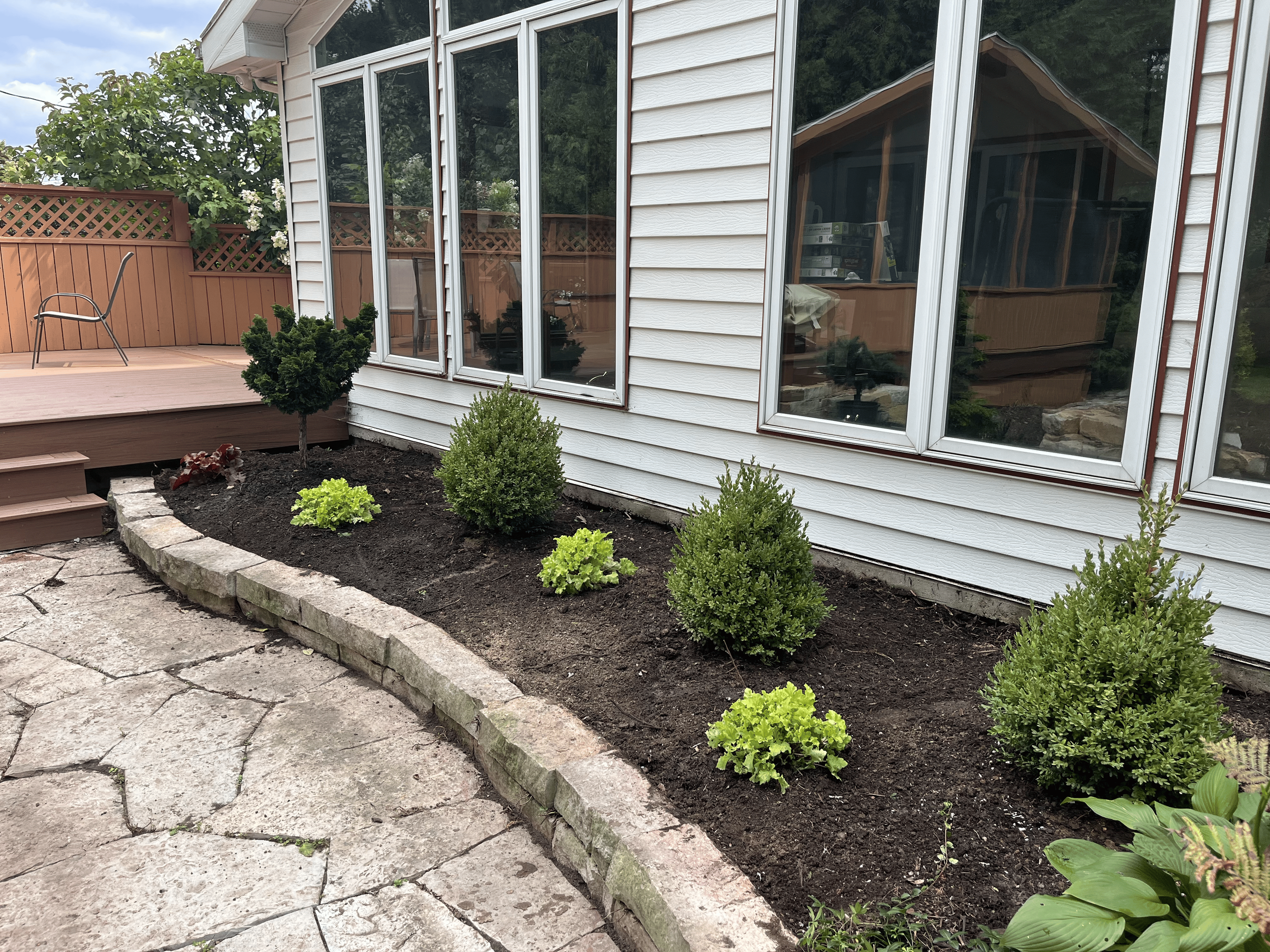 A backyard garden with a small landscaped flower bed bordered by a stone edge, containing green shrubs and plants. There is a wooden deck with a metal chair, a decorative lattice fence, and a house with white vinyl siding and large windows reflecting