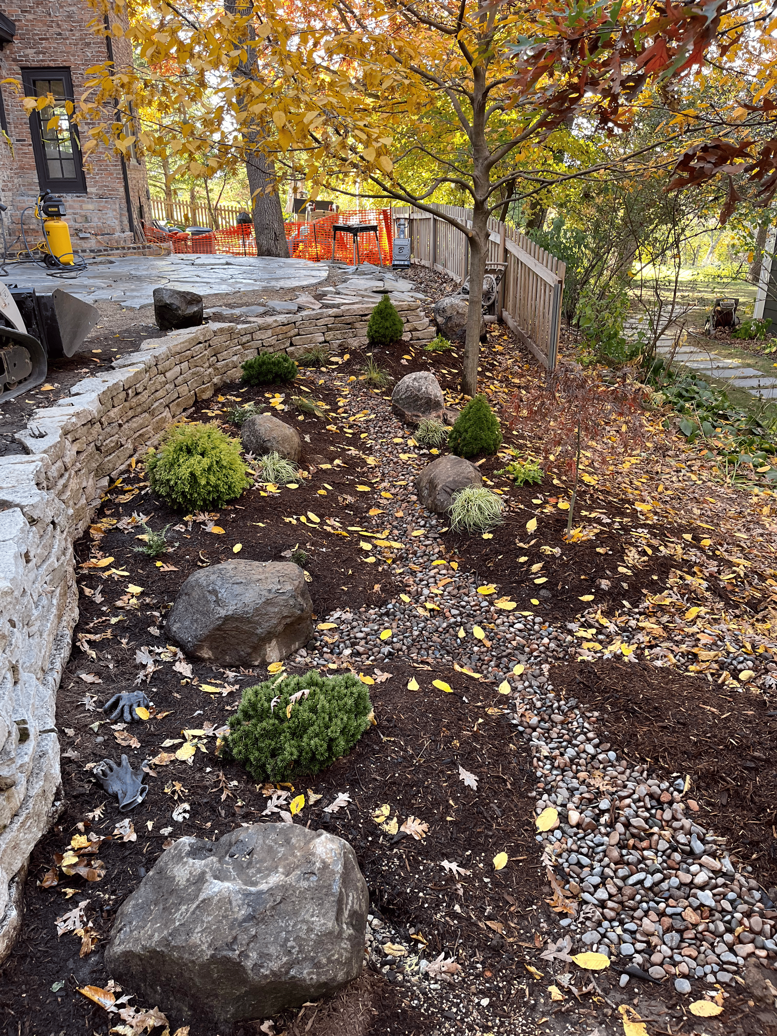 A landscaped backyard with a stone retaining wall, small shrubs, and a gravel pathway lined with larger rocks, fallen yellow leaves, a tree with autumn foliage, and a wooden fence in the background.