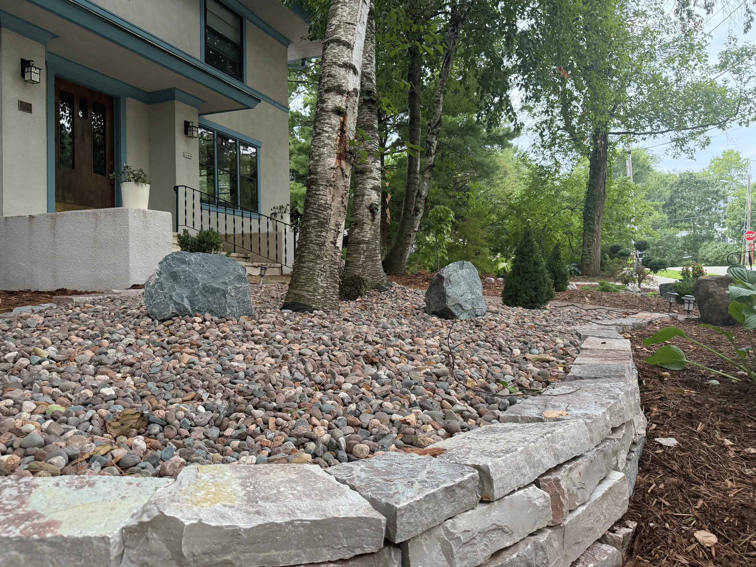 A residential front yard with trees, large rocks, pebble ground cover, and a stone pathway leading to the house entrance. The house has a modern design with a front door, large window, and outdoor lights.