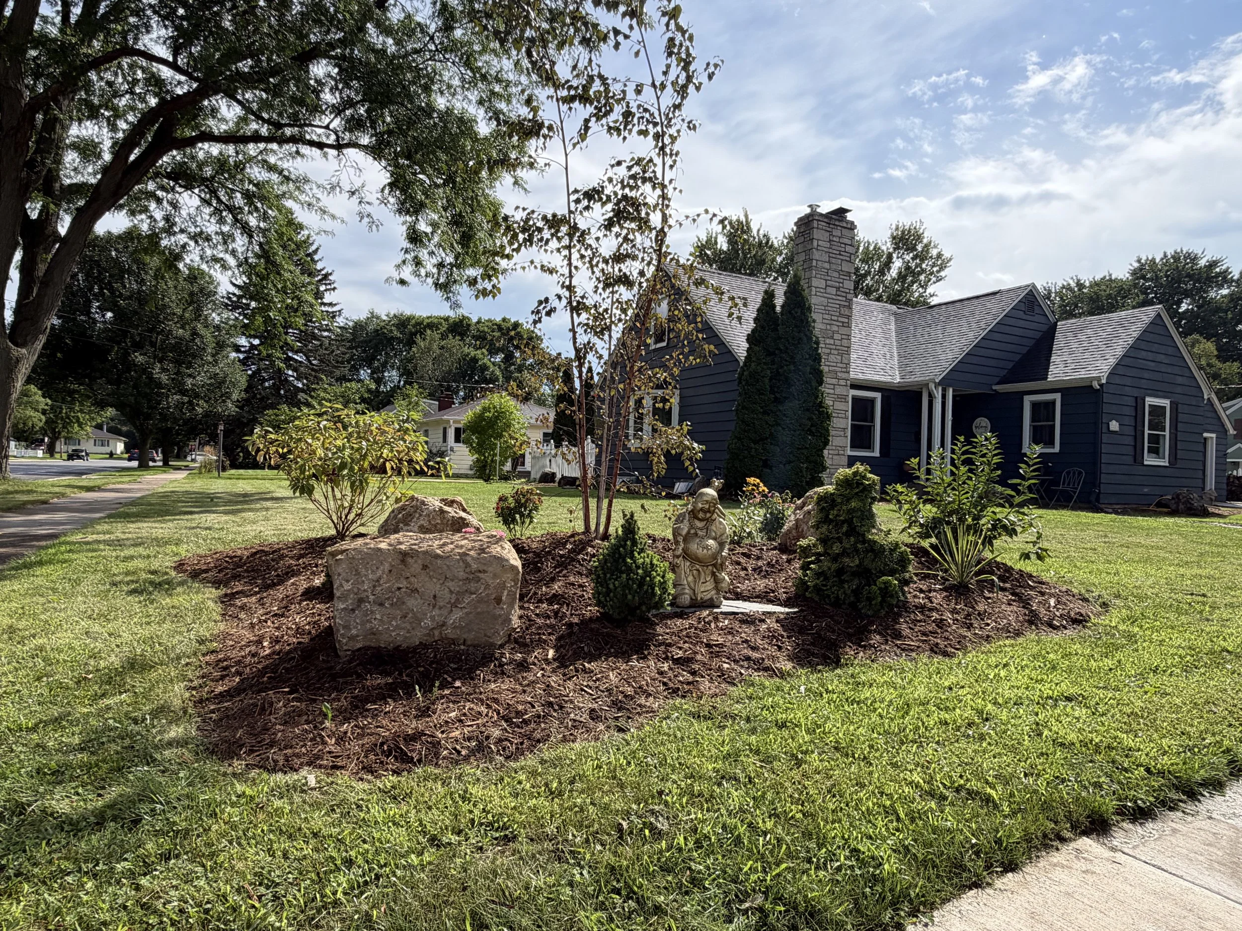 A suburban front yard with manicured grass, a garden bed with rocks, small plants, a statue of a gnome, and tall trees. A house with dark blue siding and a stone chimney is in the background, under a partly cloudy sky.