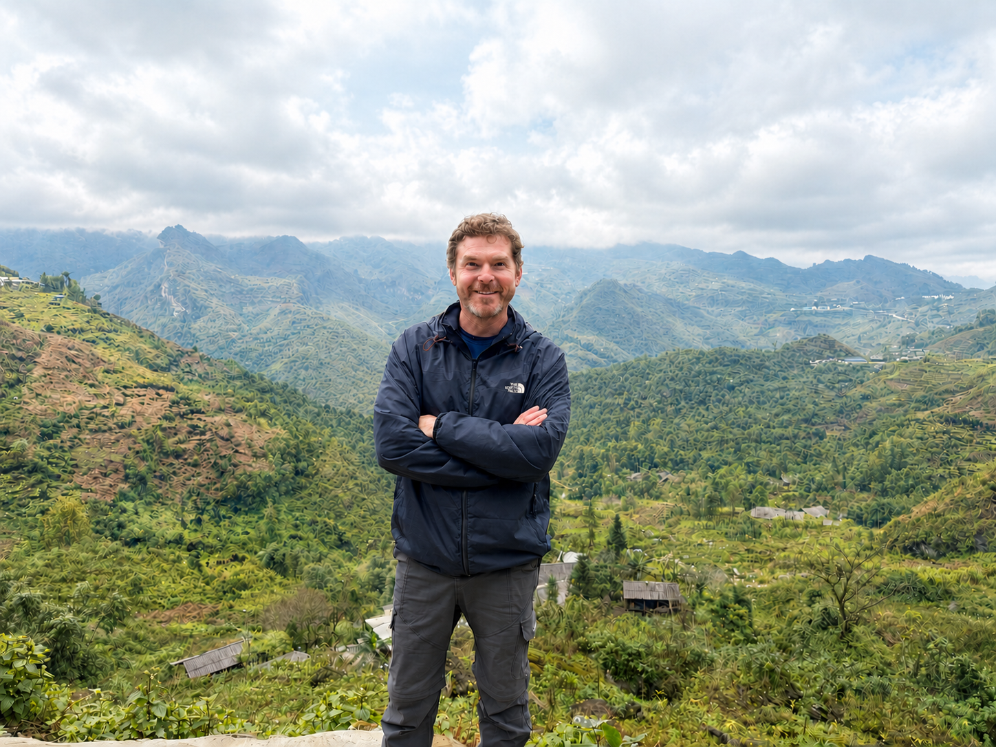 A man standing outdoors with arms crossed in front of a lush green valley and mountains, under a cloudy sky.