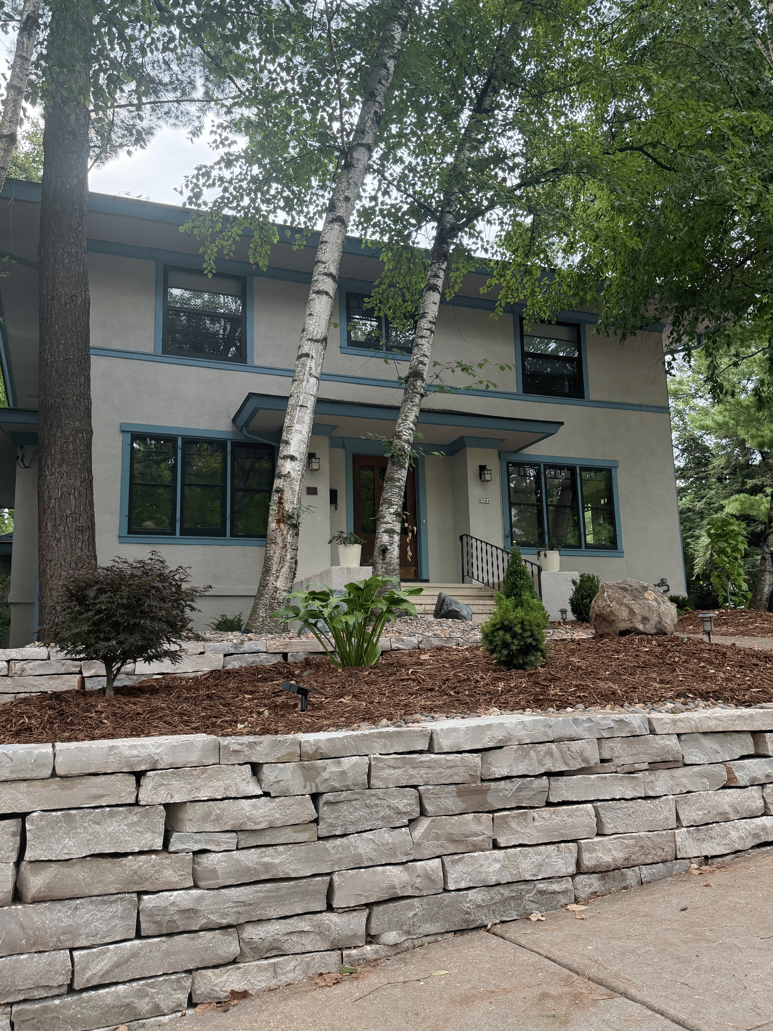 Front view of a modern two-story house with a landscaped front yard, trees, and a stone retaining wall.