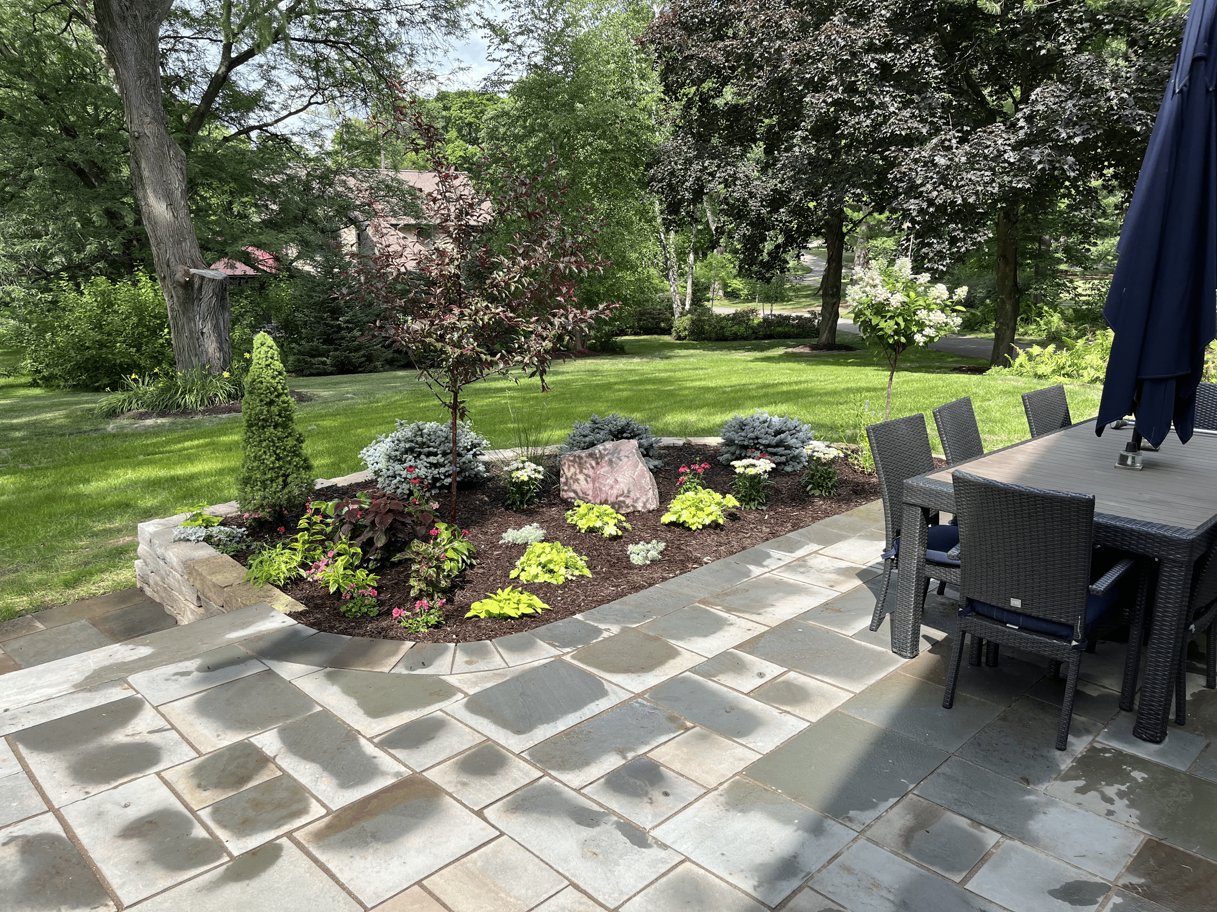 A backyard with a neatly maintained lawn and a garden bed with various flowers and shrubs. Tall trees with dense foliage surround the area. A patio with outdoor furniture, including a table and chairs, is visible on the right. A large dark blue umbre